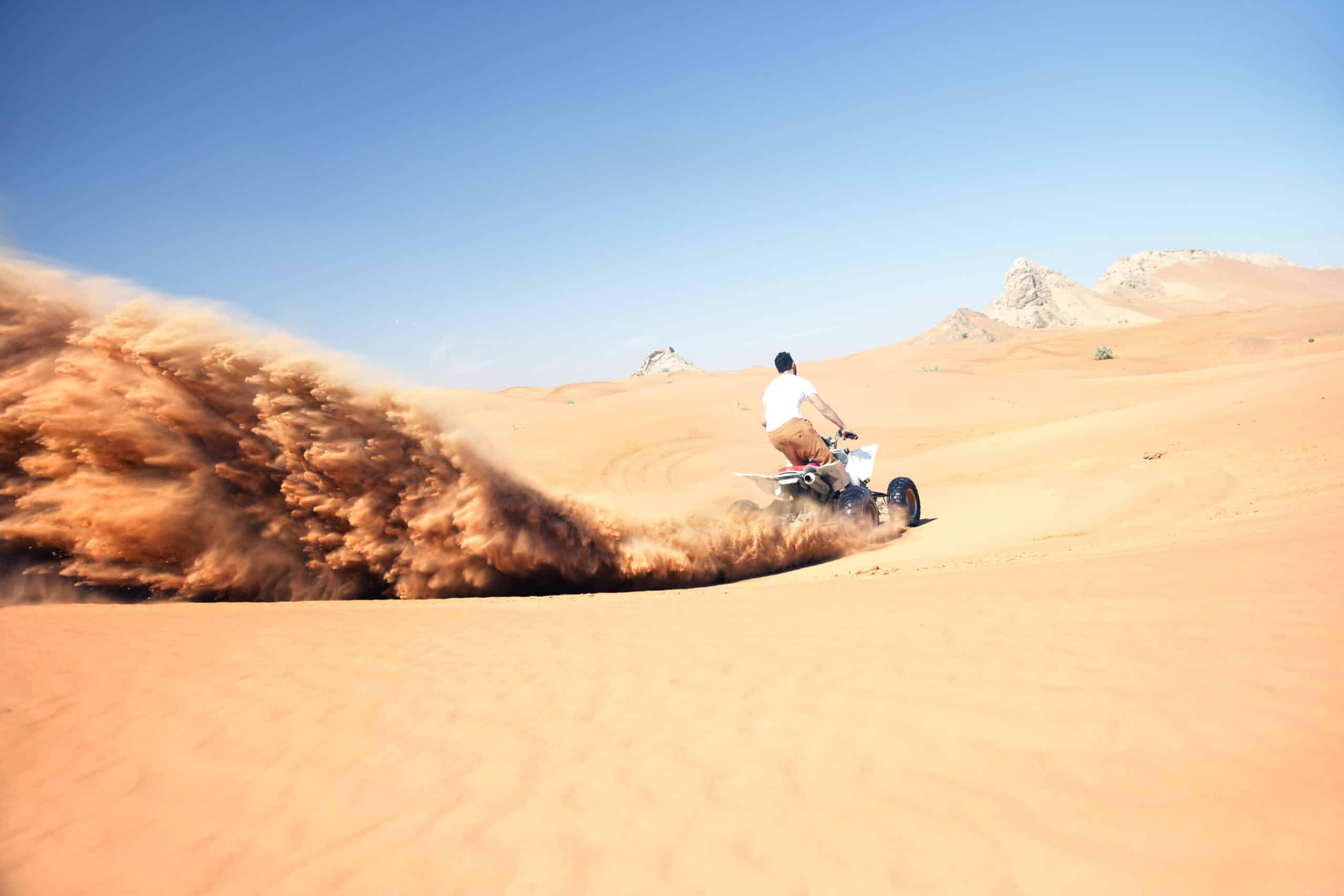 Eine Person fährt mit hoher Geschwindigkeit auf einem Motorrad durch eine Sandwüste und wirbelt dabei eine große Sandspur auf. Im Hintergrund sind unter einem klaren blauen Himmel Berge zu sehen. © Fotografie Tomas Rodriguez