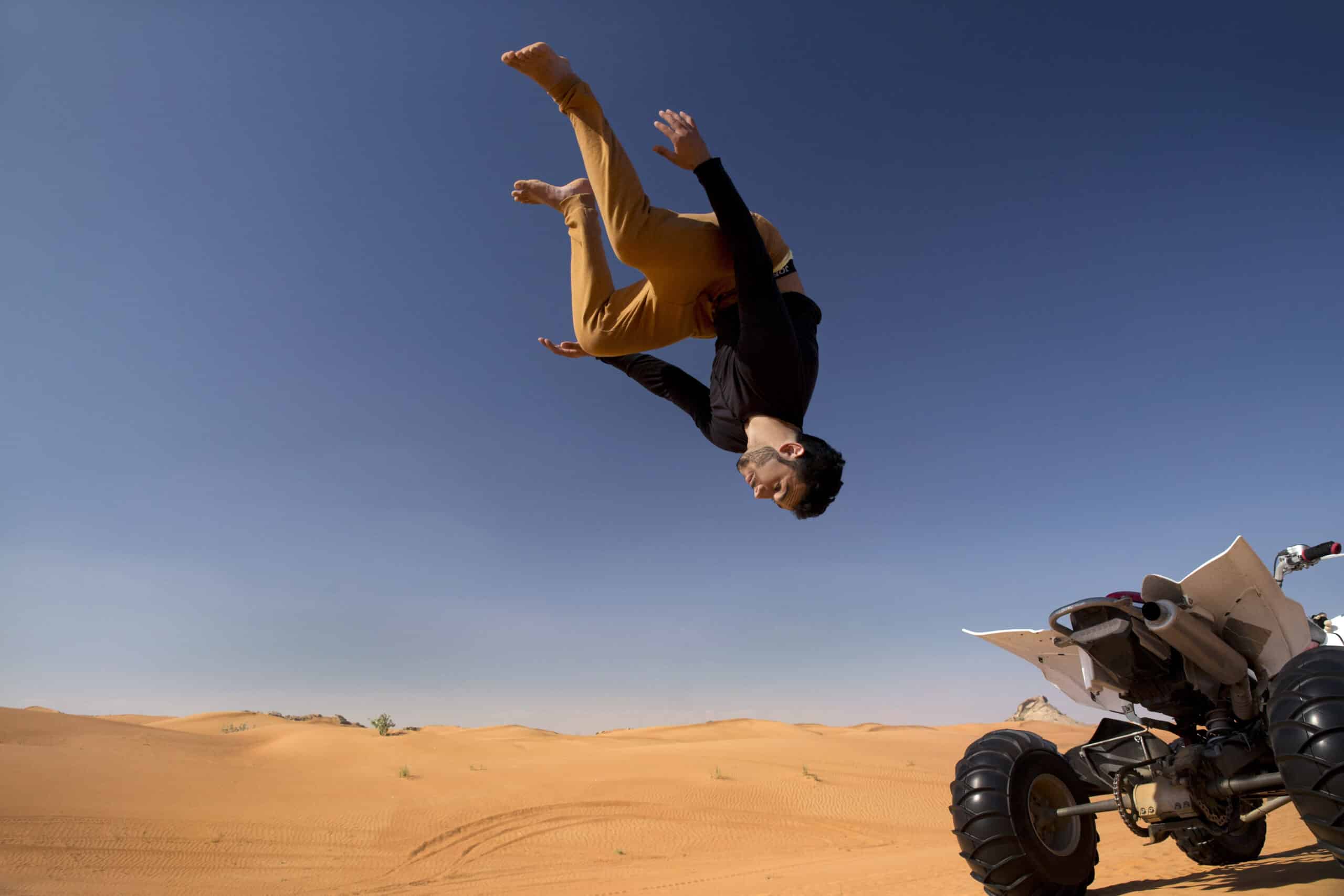 Eine Person in einem gelben Oberteil und einer schwarzen Hose vollführt in einer riesigen Wüstenlandschaft unter einem klaren blauen Himmel einen akrobatischen Saltos über einem Quad. © Fotografie Tomas Rodriguez