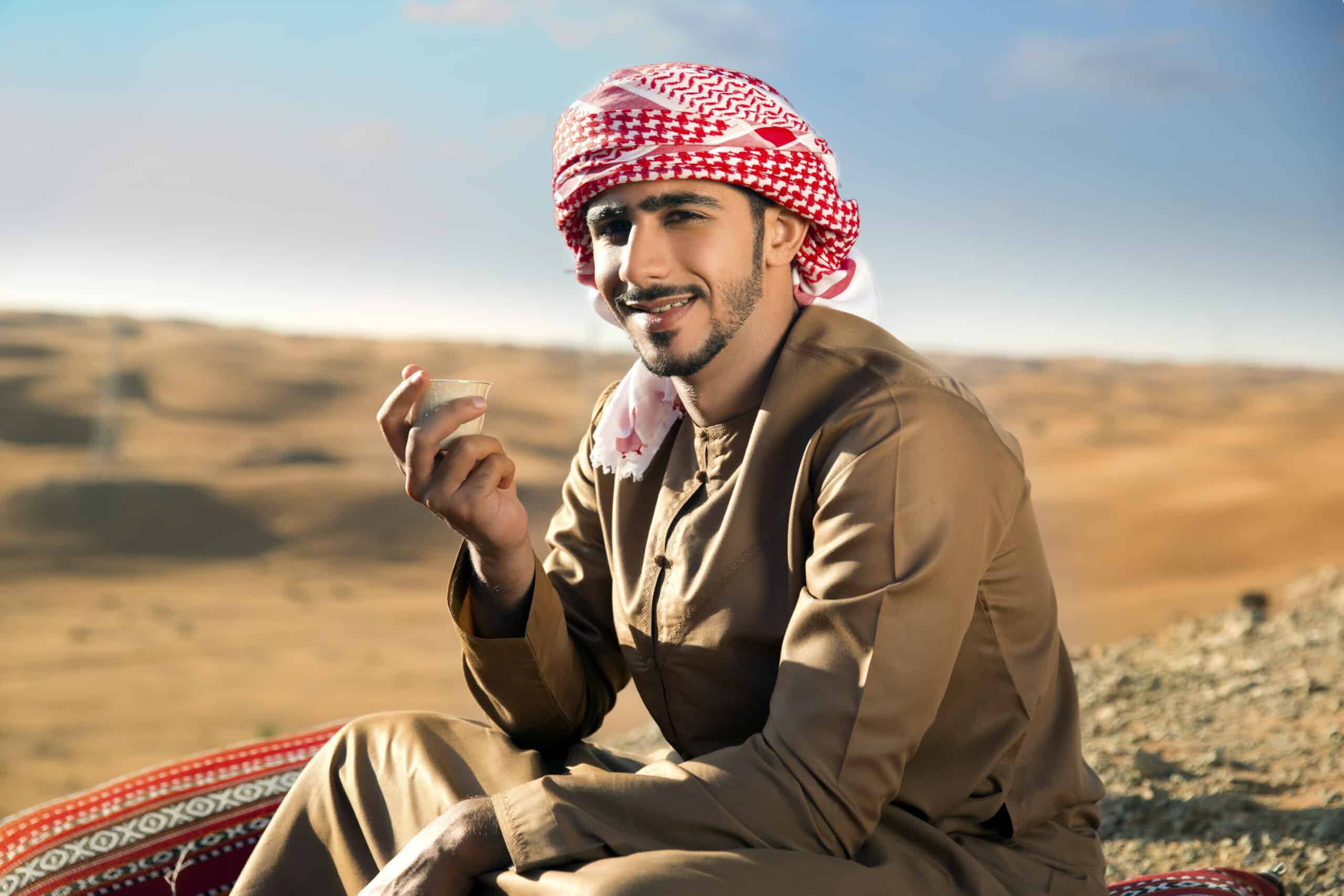 Ein lächelnder Mann in traditioneller nahöstlicher Kleidung sitzt auf einem gemusterten Teppich in einer Wüstenlandschaft und hält eine kleine Tasse Kaffee. © Fotografie Tomas Rodriguez