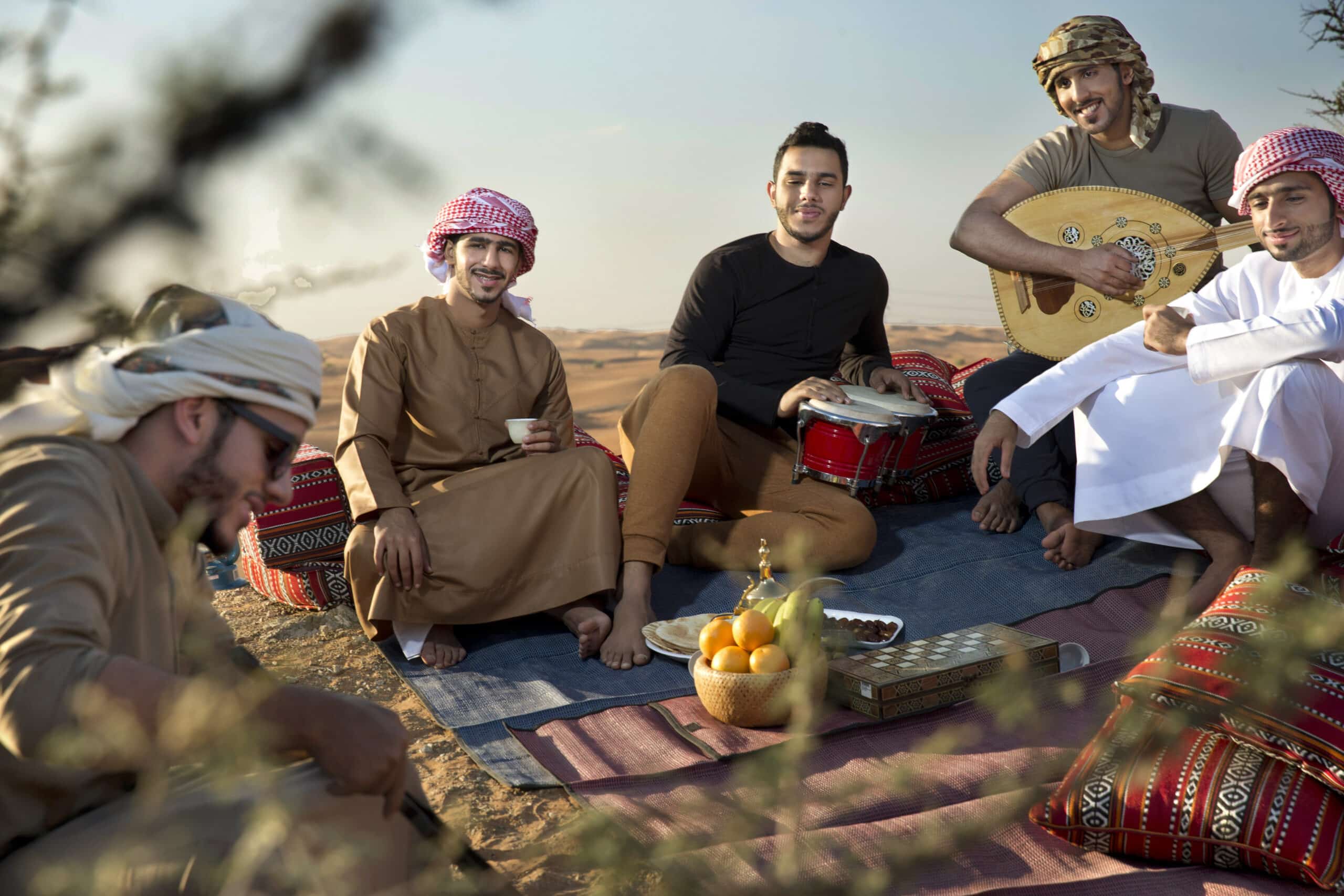 Eine Gruppe von fünf Männern in traditioneller nahöstlicher Kleidung sitzt auf einem bunten Teppich in einer Wüstenlandschaft und genießt die Gesellschaft des anderen, während einer von ihnen Trommel und ein anderer ein Saiteninstrument spielt. © Fotografie Tomas Rodriguez