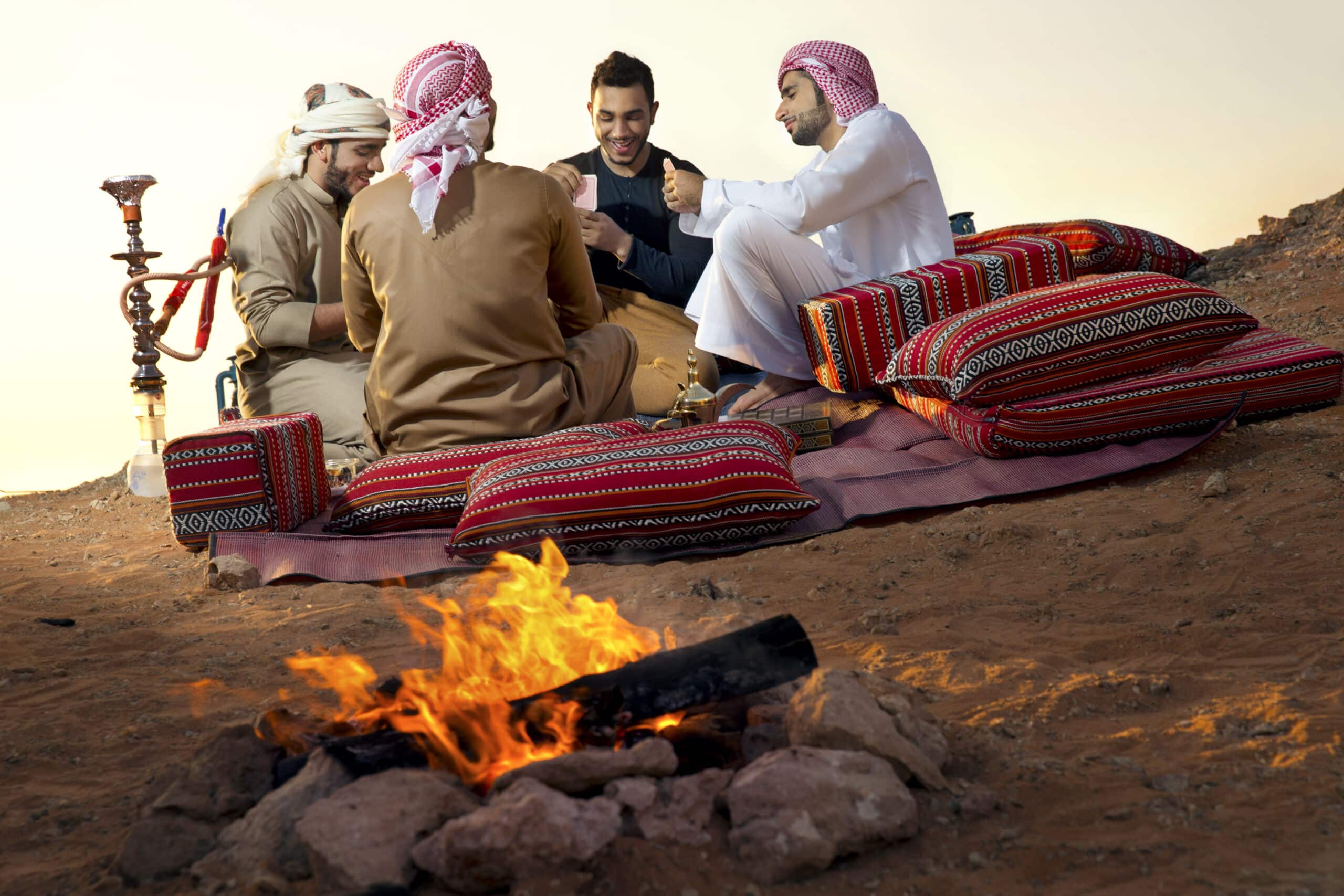 Eine Gruppe von Männern in traditioneller nahöstlicher Kleidung sitzt auf bunten Kissen in einer Wüste und unterhält sich an einem kleinen Lagerfeuer. In der Nähe steht eine Wasserpfeife. © Fotografie Tomas Rodriguez