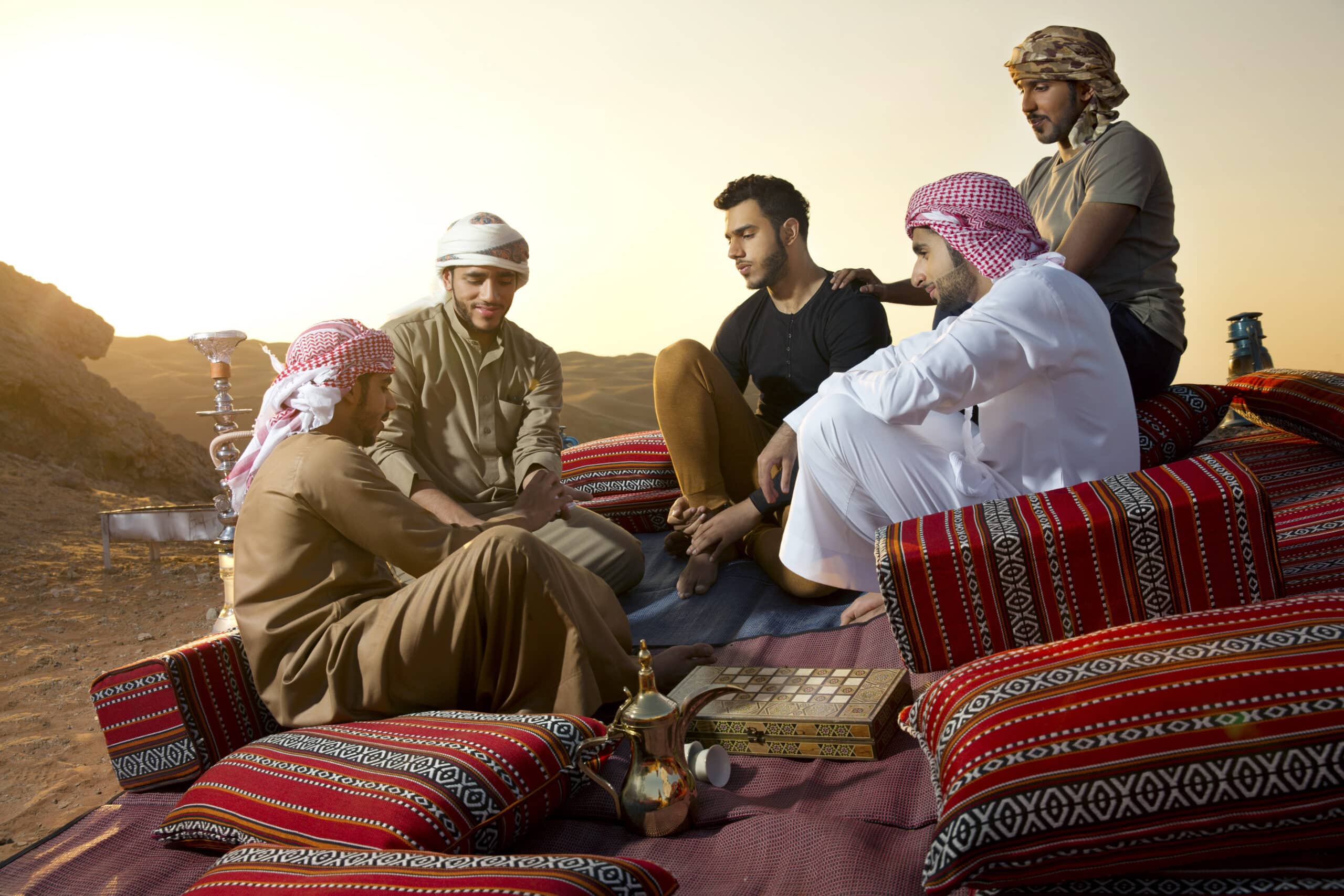 Eine Gruppe von fünf Männern in traditioneller nahöstlicher Kleidung sitzt bei Sonnenuntergang auf verzierten Teppichen in einer Wüste und unterhält sich vor einer Teekanne und Tassen. © Fotografie Tomas Rodriguez