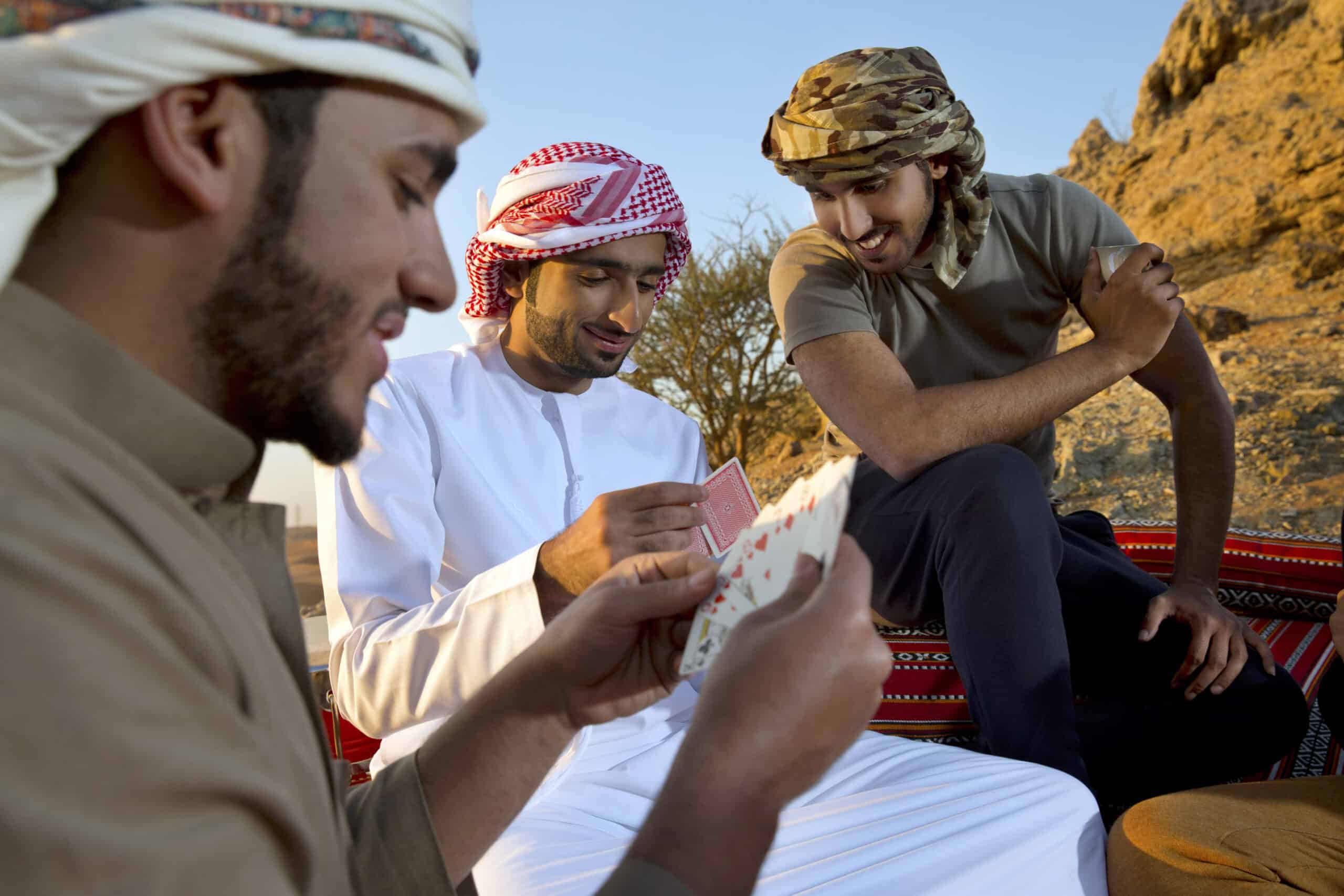 Drei Männer in traditioneller arabischer Kleidung sitzen im Freien, spielen Karten und lächeln, mit felsigem Gelände im Hintergrund bei Sonnenuntergang. © Fotografie Tomas Rodriguez