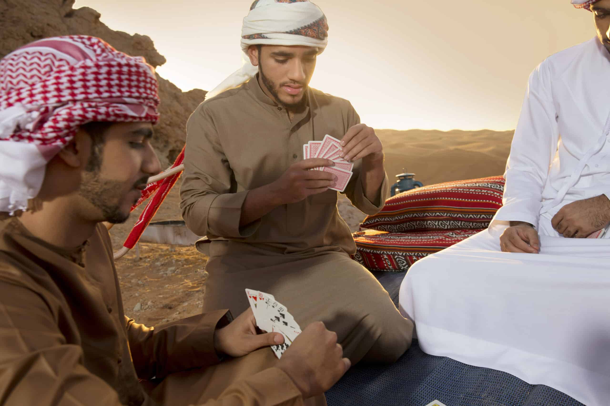 Drei Männer in traditioneller nahöstlicher Kleidung sitzen auf Kissen in einer Wüste und spielen Karten. Ein Mann konzentriert sich auf seine Hand, während die anderen zuschauen. © Fotografie Tomas Rodriguez