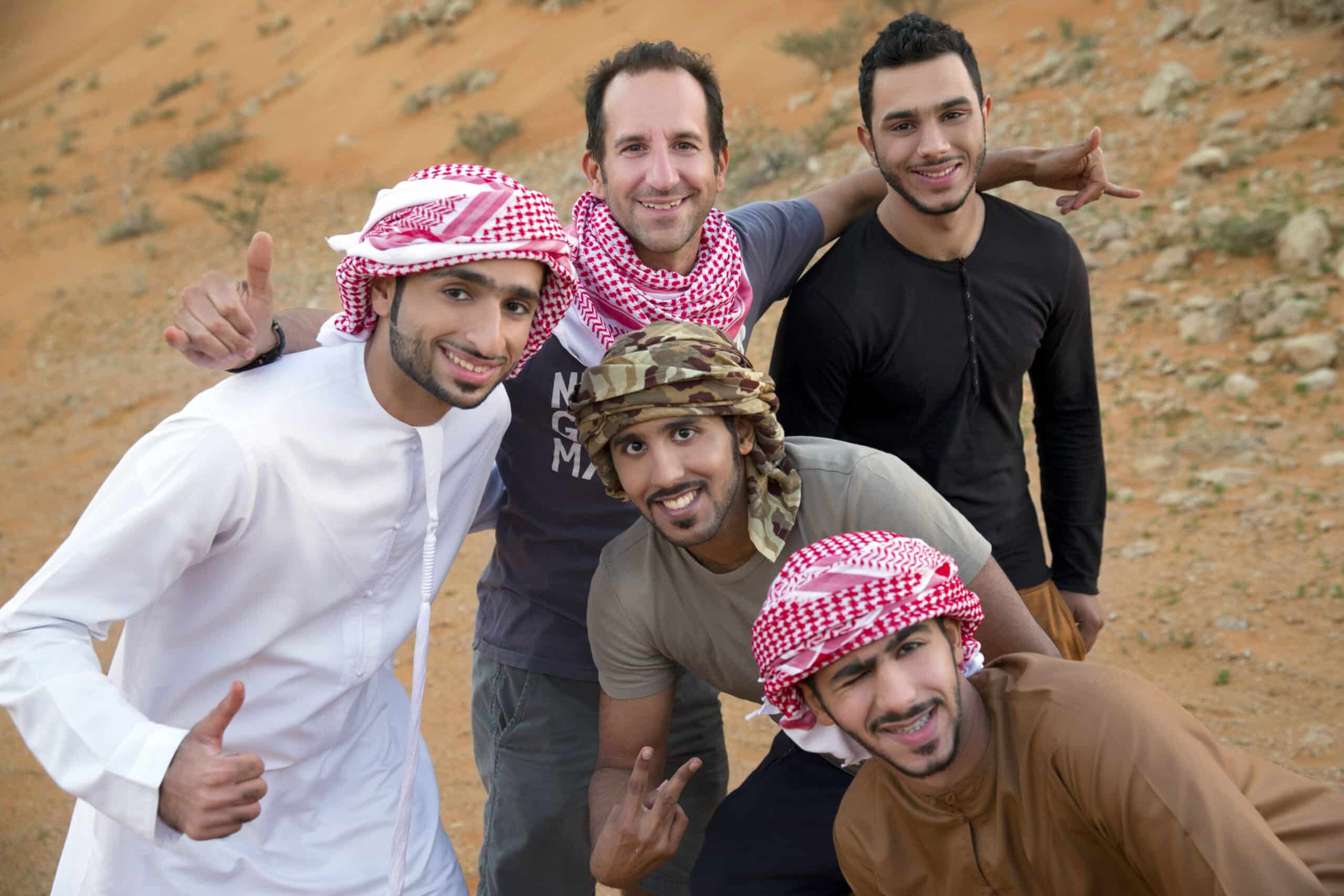 Fünf Männer in einer Wüstenlandschaft, vier tragen traditionelle Kufiyas und einer in westlicher Kleidung, und posieren fröhlich mit einer freundlichen Geste. © Fotografie Tomas Rodriguez