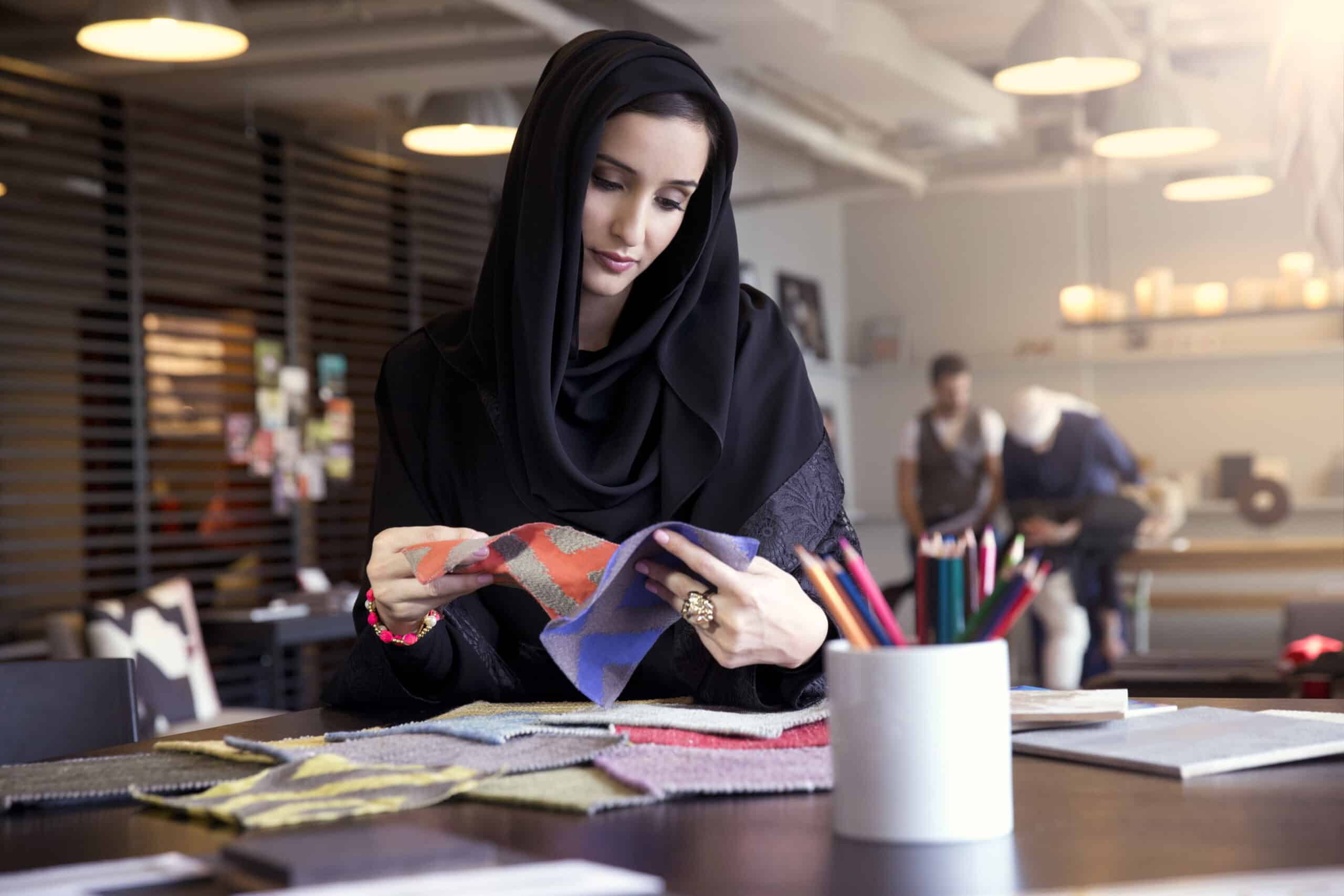 Eine Frau mit Hijab untersucht Stoffmuster an einem Tisch in einem hellen, modernen Büro, während im Hintergrund Kollegen arbeiten. © Fotografie Tomas Rodriguez