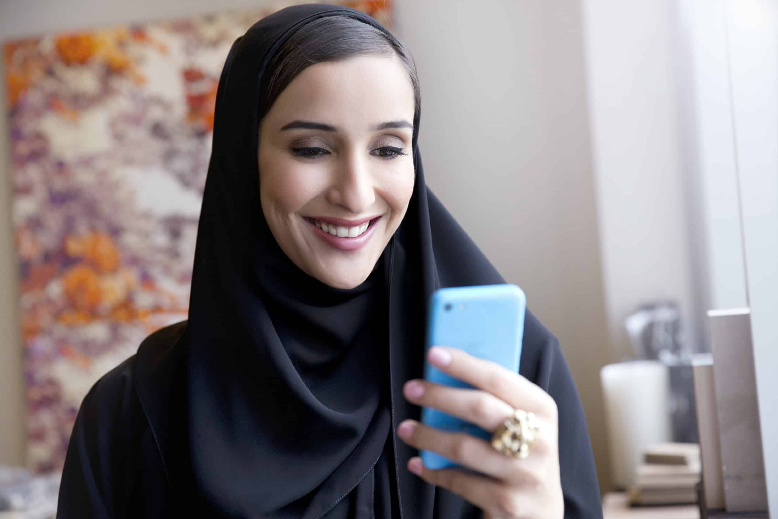 Eine Frau im Hijab lächelt fröhlich, während sie in einem gut beleuchteten Raum mit floralen Kunstwerken im Hintergrund auf ihr blaues Smartphone schaut. © Fotografie Tomas Rodriguez