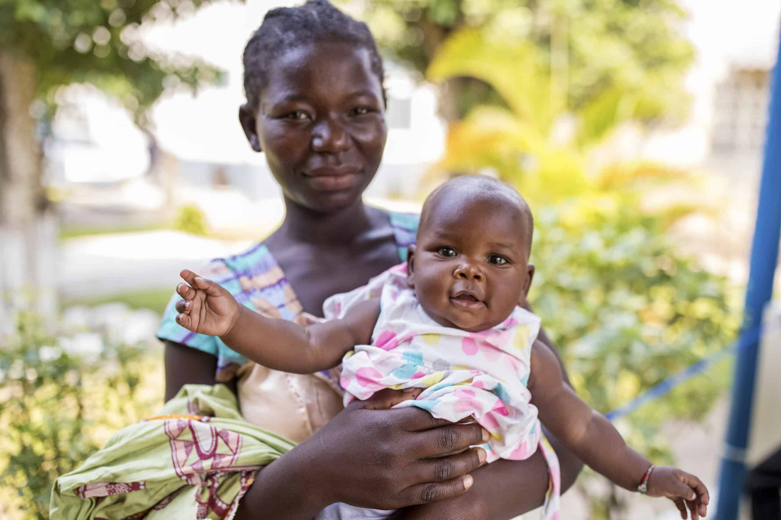 Eine junge Afrikanerin hält ein lächelndes Baby auf einem Arm. Sie sind im Freien, im Hintergrund sind verschwommene grüne Bäume zu sehen. Das Baby trägt ein rosa-weißes Outfit und die Frau blickt in die Kamera. © Fotografie Tomas Rodriguez