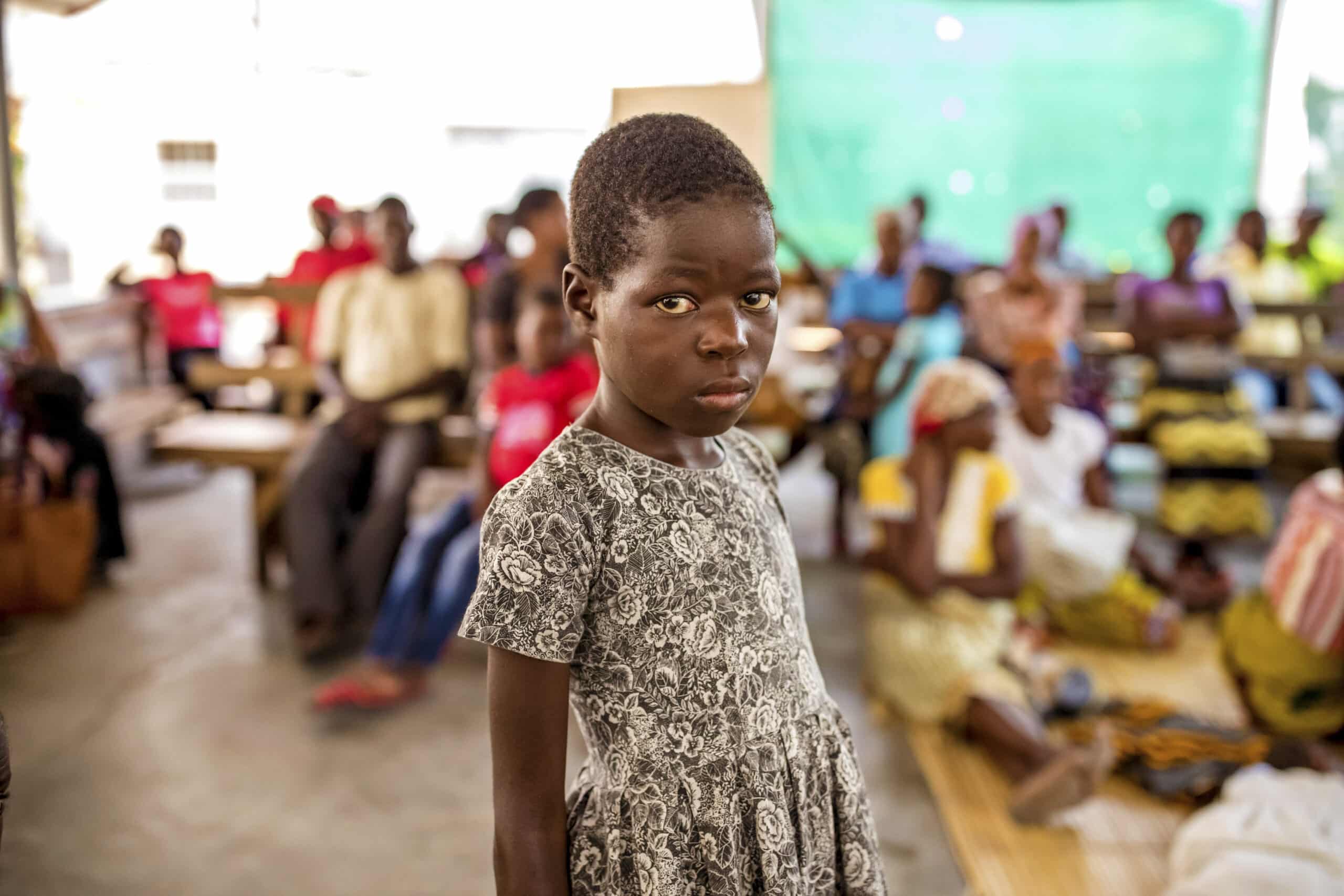 Im Fokus steht ein junges afrikanisches Mädchen, das ernst in die Kamera blickt. Im Hintergrund sind unscharfe Menschen zu sehen, die in einer zeltartigen Konstruktion sitzen. © Fotografie Tomas Rodriguez