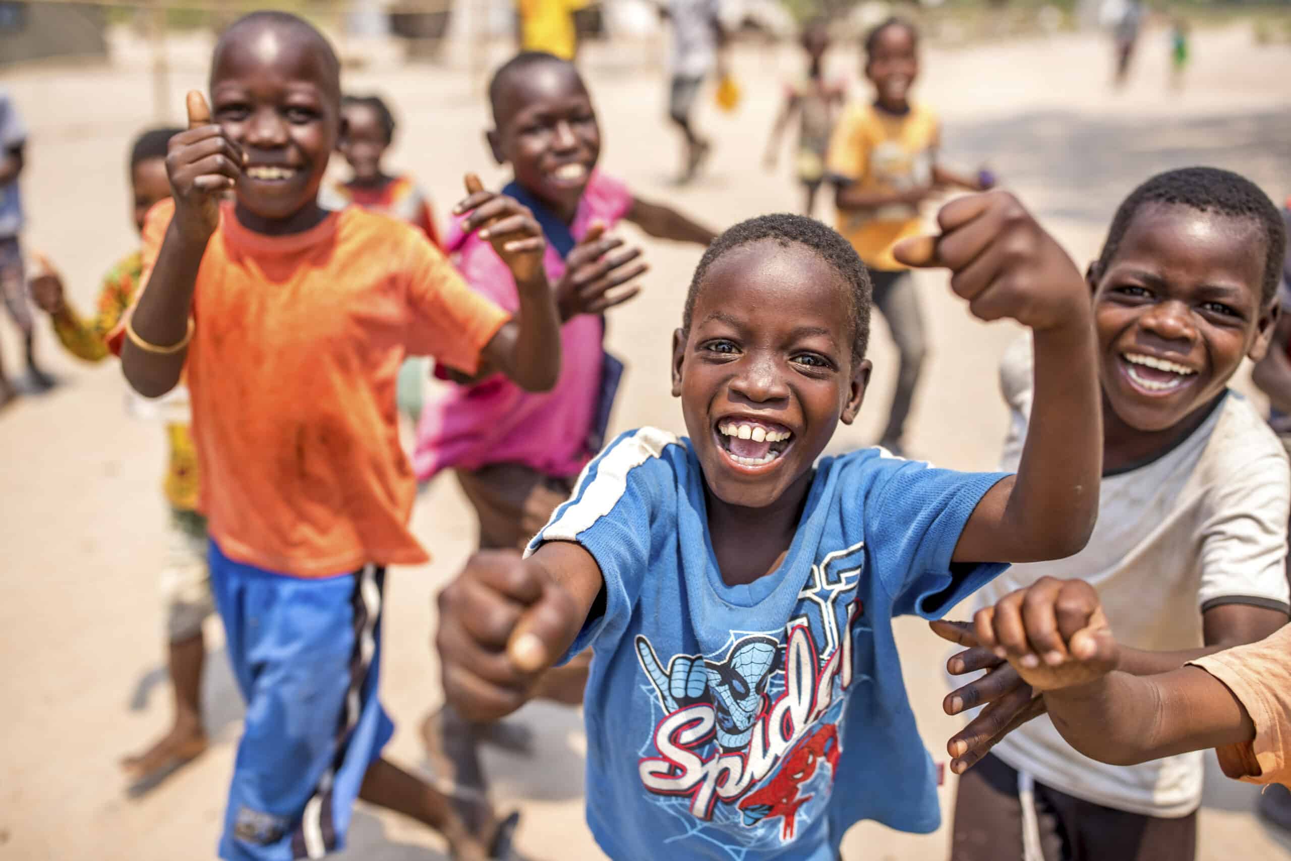Eine Gruppe fröhlicher afrikanischer Kinder rennt an einem sonnigen Tag in einem sandigen Gebiet lächelnd auf die Kamera zu und gestikuliert mit dem Daumen nach oben. © Fotografie Tomas Rodriguez