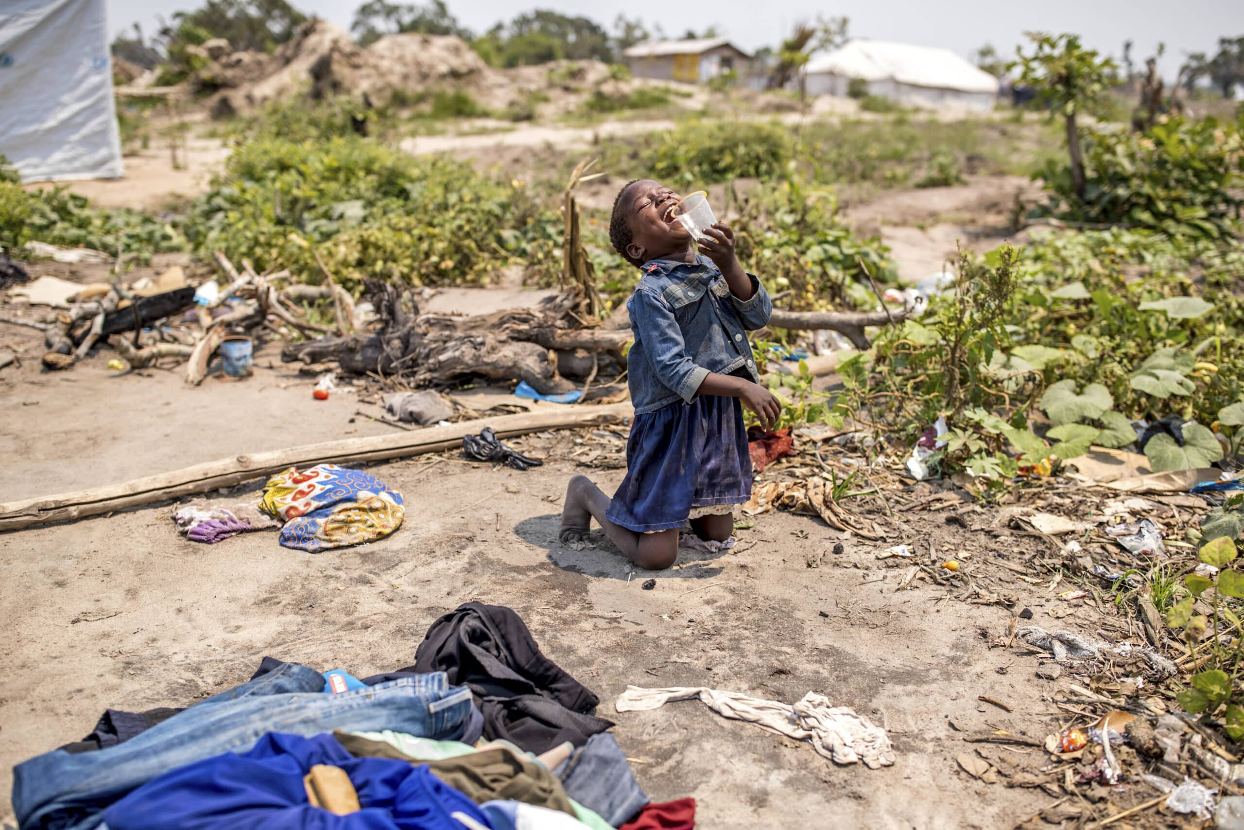 Ein junges Mädchen kniet in einem provisorischen Lager auf dem Boden und weint mit der Hand vor dem Mund. Um sie herum liegen verstreute Kleidungsstücke und Trümmer, im Hintergrund sind Zelte und eine öde Landschaft zu sehen. © Fotografie Tomas Rodriguez