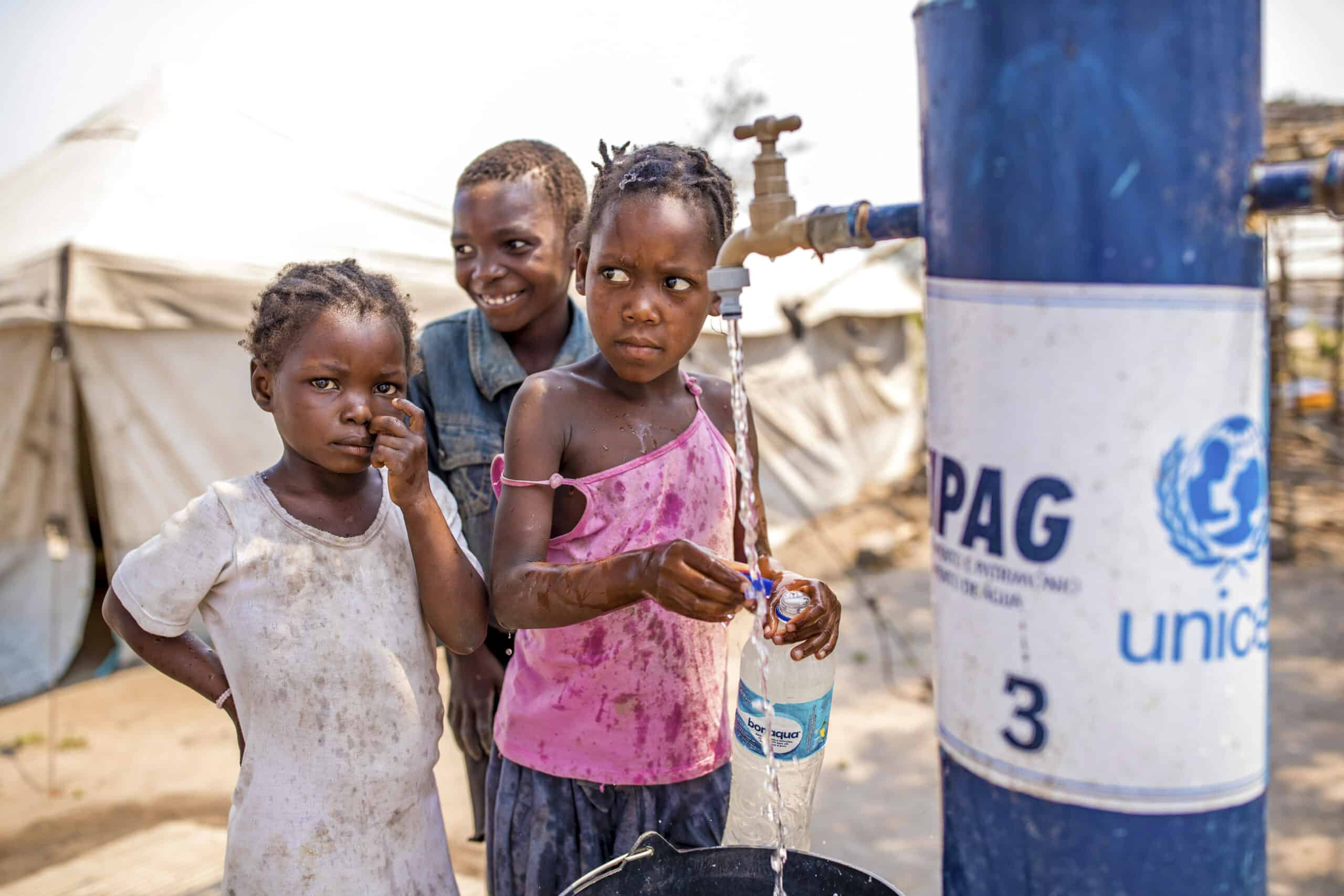 Drei Kinder an einer Wasserstelle in einem Flüchtlingslager. Ein junges Mädchen füllt eine Flasche, ein anderes Mädchen steht daneben und im Hintergrund lächelt ein Junge. Im Hintergrund sind Zelte und trockene Vegetation zu sehen. © Fotografie Tomas Rodriguez