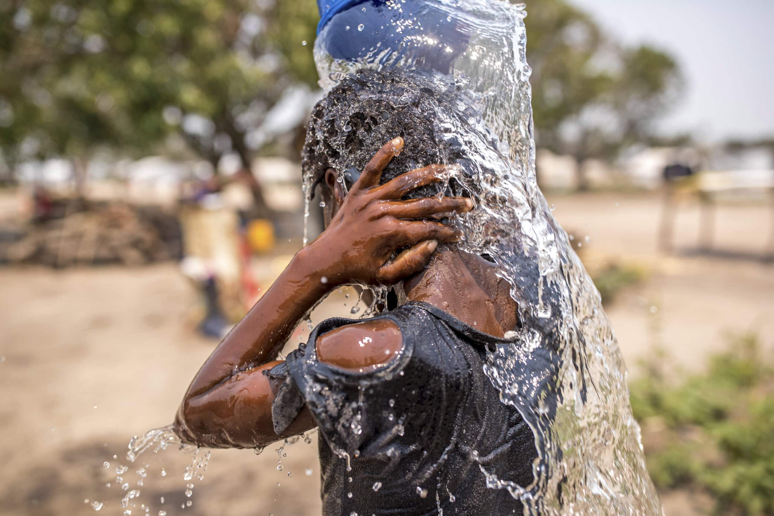 Ein kleiner Junge in einem blauen Hemd spritzt sich an einem sonnigen Tag im Freien Wasser aus einem blauen Eimer über den Kopf und reinigt sein Gesicht. Im Hintergrund sind verschwommene Bäume zu sehen. © Fotografie Tomas Rodriguez