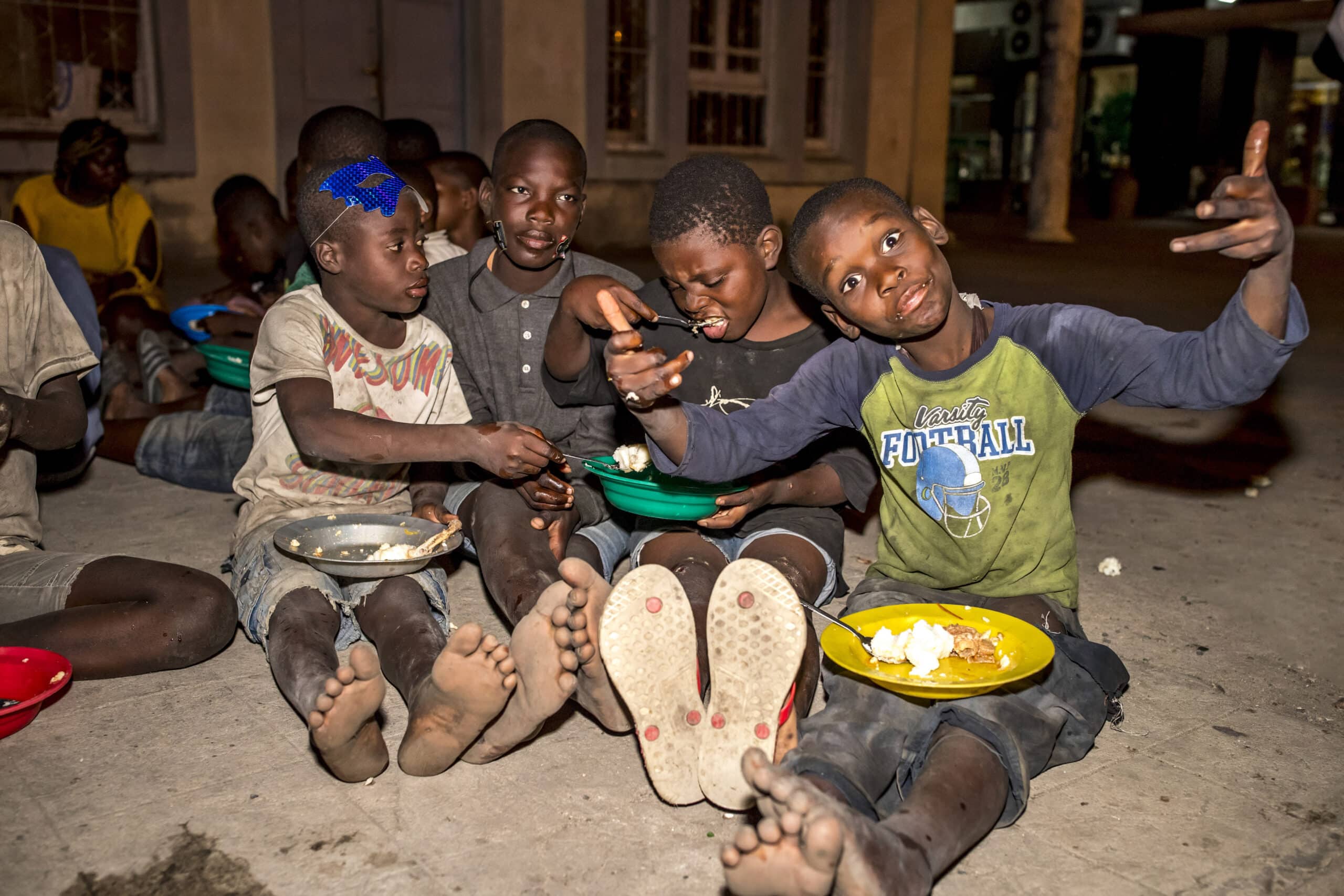 Eine Gruppe von sechs Jungen sitzt abends auf dem Boden und isst gemeinsam. Sie lächeln und unterhalten sich miteinander, vor ihnen stehen Teller mit Essen. © Fotografie Tomas Rodriguez