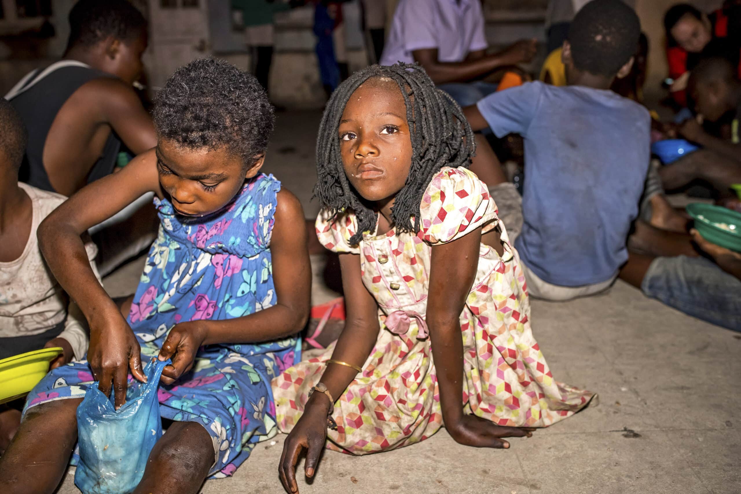 Zwei junge Mädchen sitzen nachts auf dem Boden; eines ist darauf konzentriert, ein blaues Stück Stoff zu bemalen, während das andere direkt in die Kamera blickt. Im Hintergrund sind weitere Kinder zu sehen. © Fotografie Tomas Rodriguez