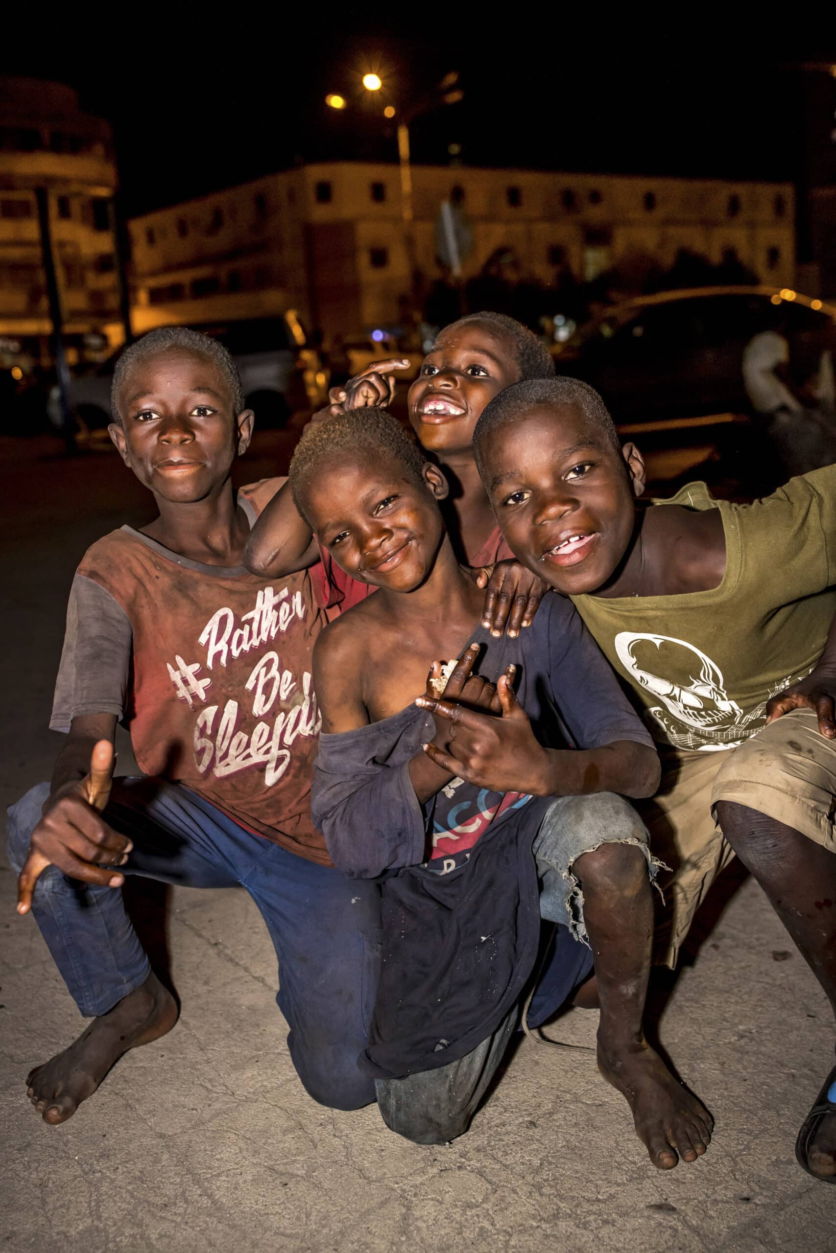 Vier lächelnde Jungen drängen sich nachts auf einer Stadtstraße zusammen. Sie wirken fröhlich und verspielt, wobei einer von ihnen das Peace-Zeichen macht. Sie tragen abgetragene Kleidung, was ein Gefühl der Kameradschaft ausdrückt. © Fotografie Tomas Rodriguez