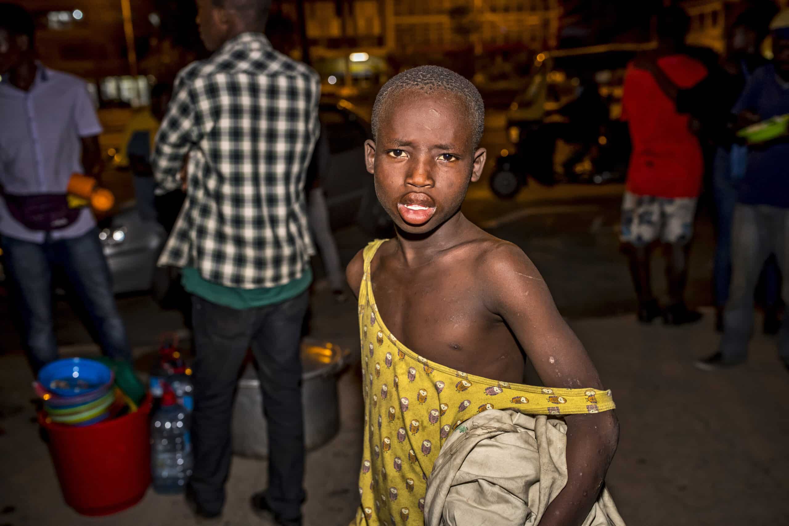 Ein kleiner Junge in einem ärmellosen Oberteil steht überrascht in einer nächtlichen Straßenszenerie. Im unscharfen Hintergrund sind Menschen und Straßenhändler zu sehen. © Fotografie Tomas Rodriguez