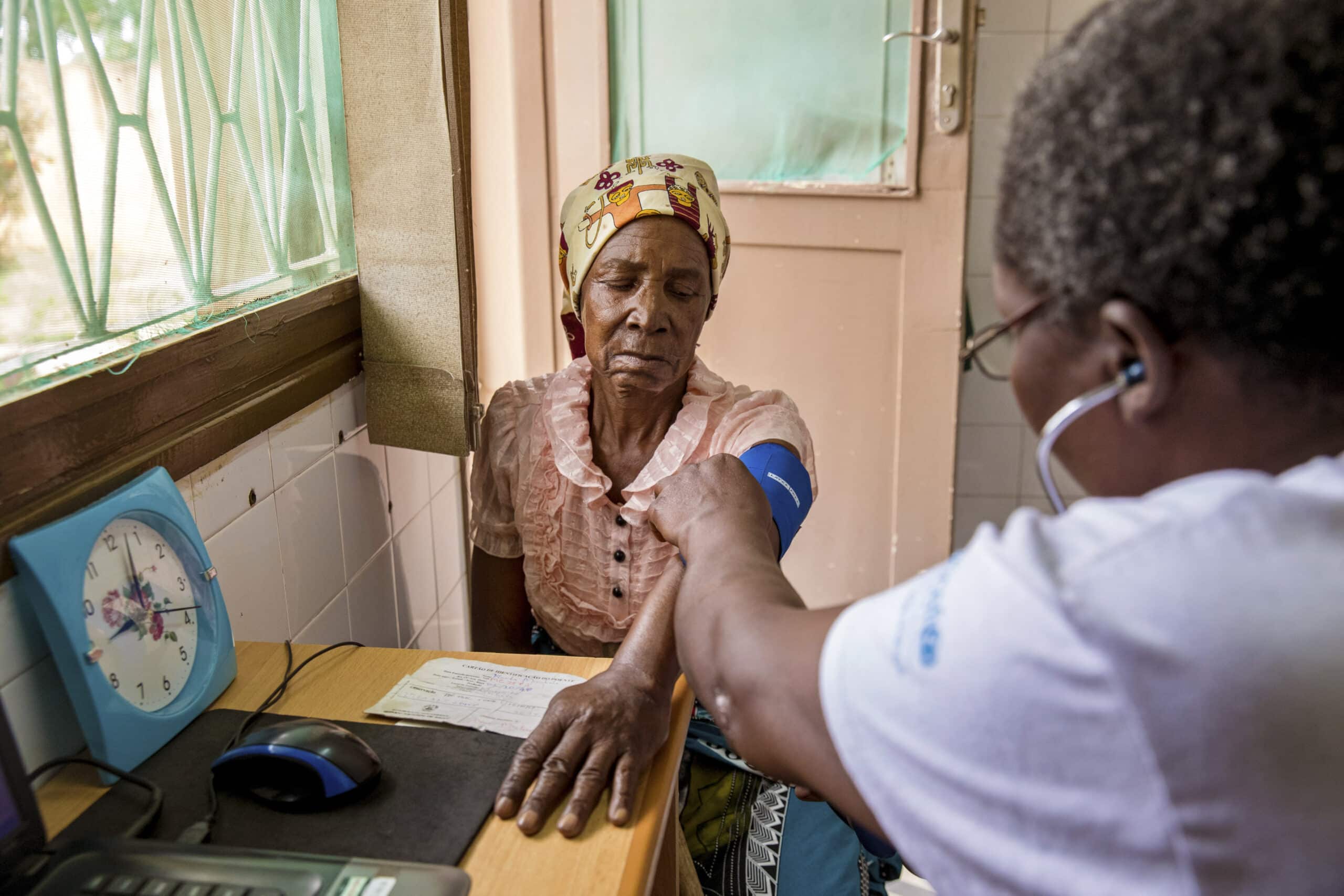 Ein Mitarbeiter des Gesundheitswesens misst den Blutdruck einer älteren Frau in einer Klinik, im Hintergrund sind eine Uhr und ein Fenster zu sehen. © Fotografie Tomas Rodriguez