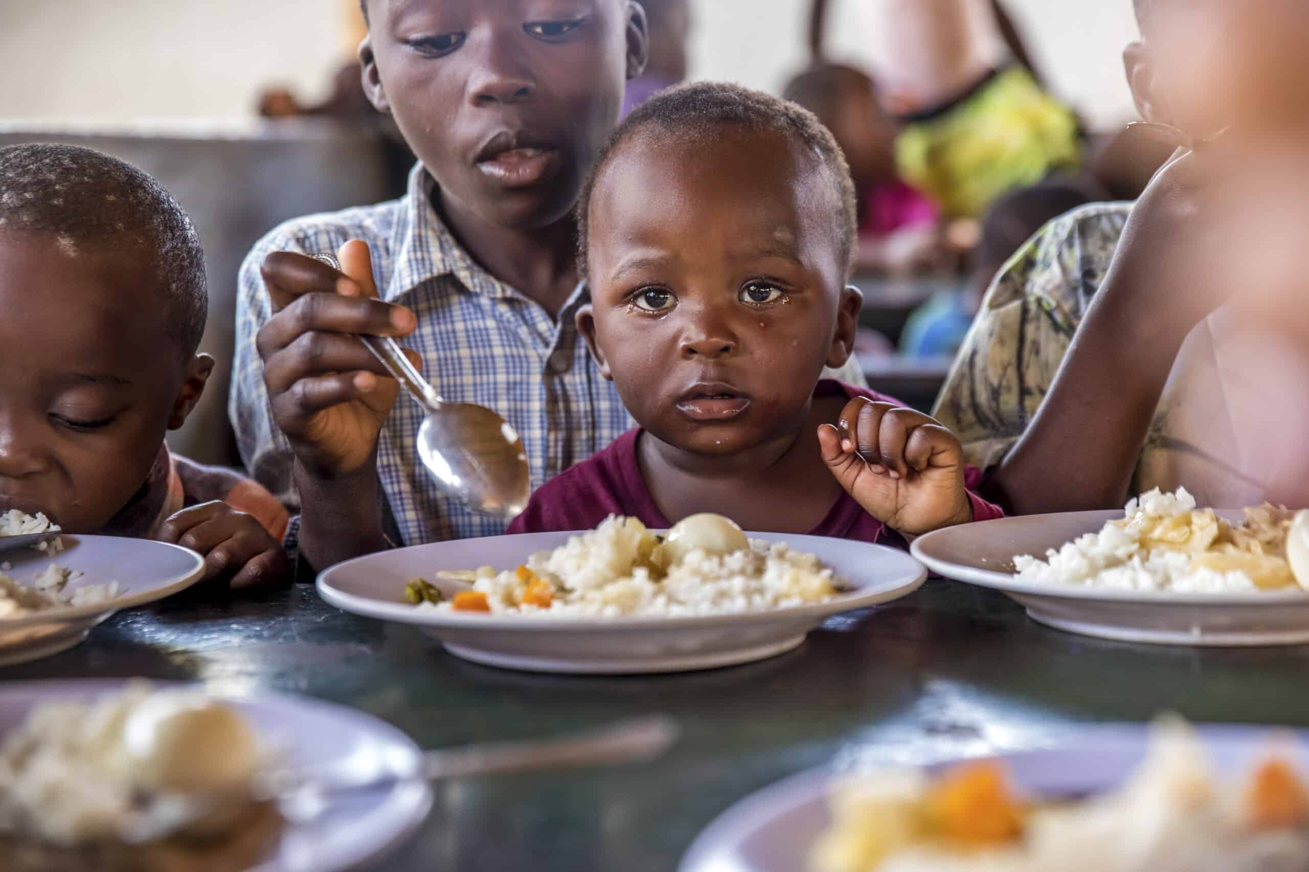 Kleine Kinder essen gemeinsam an einem Tisch, vor jedem steht ein Teller Reis und Gemüse; ein Kleinkind blickt direkt in die Kamera. © Fotografie Tomas Rodriguez