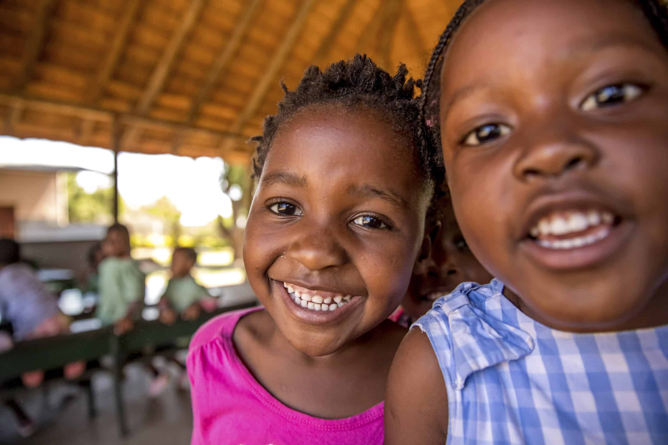 Zwei fröhliche junge Mädchen lächeln in die Kamera. Eines trägt ein rosa Oberteil und das andere ein blau-weiß gestreiftes Oberteil. Sie befinden sich in einem Holzpavillon mit den anderen Mädchen im Hintergrund. © Fotografie Tomas Rodriguez