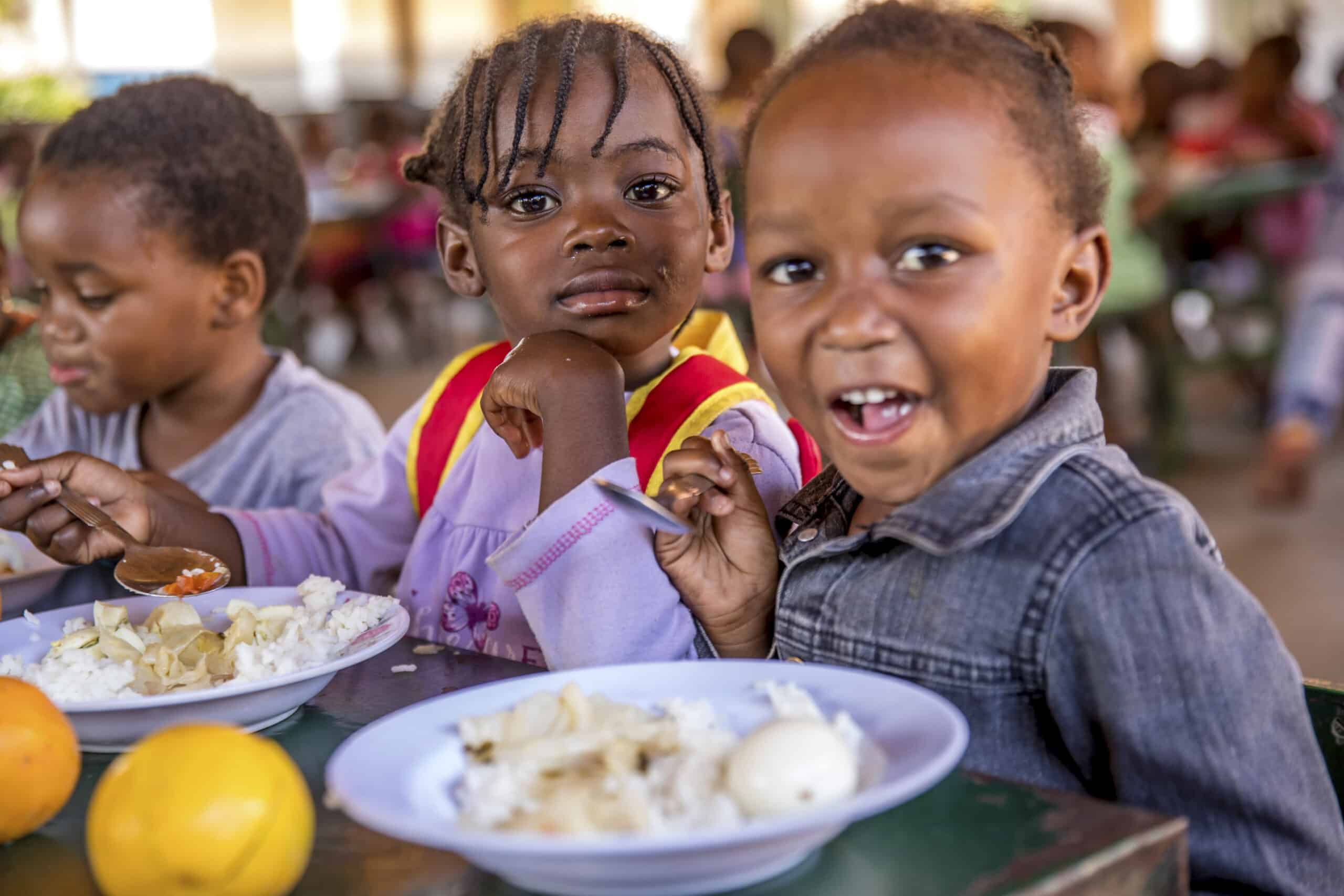 Drei kleine Kinder essen an einem Tisch. Zwei blicken in die Kamera – ein breit lächelnder Junge und ein neugierig dreinblickendes Mädchen mit geflochtenen Haaren. Vor ihnen stehen Teller mit Essen und Obst in einer hellen Cafeteria-Umgebung. © Fotografie Tomas Rodriguez