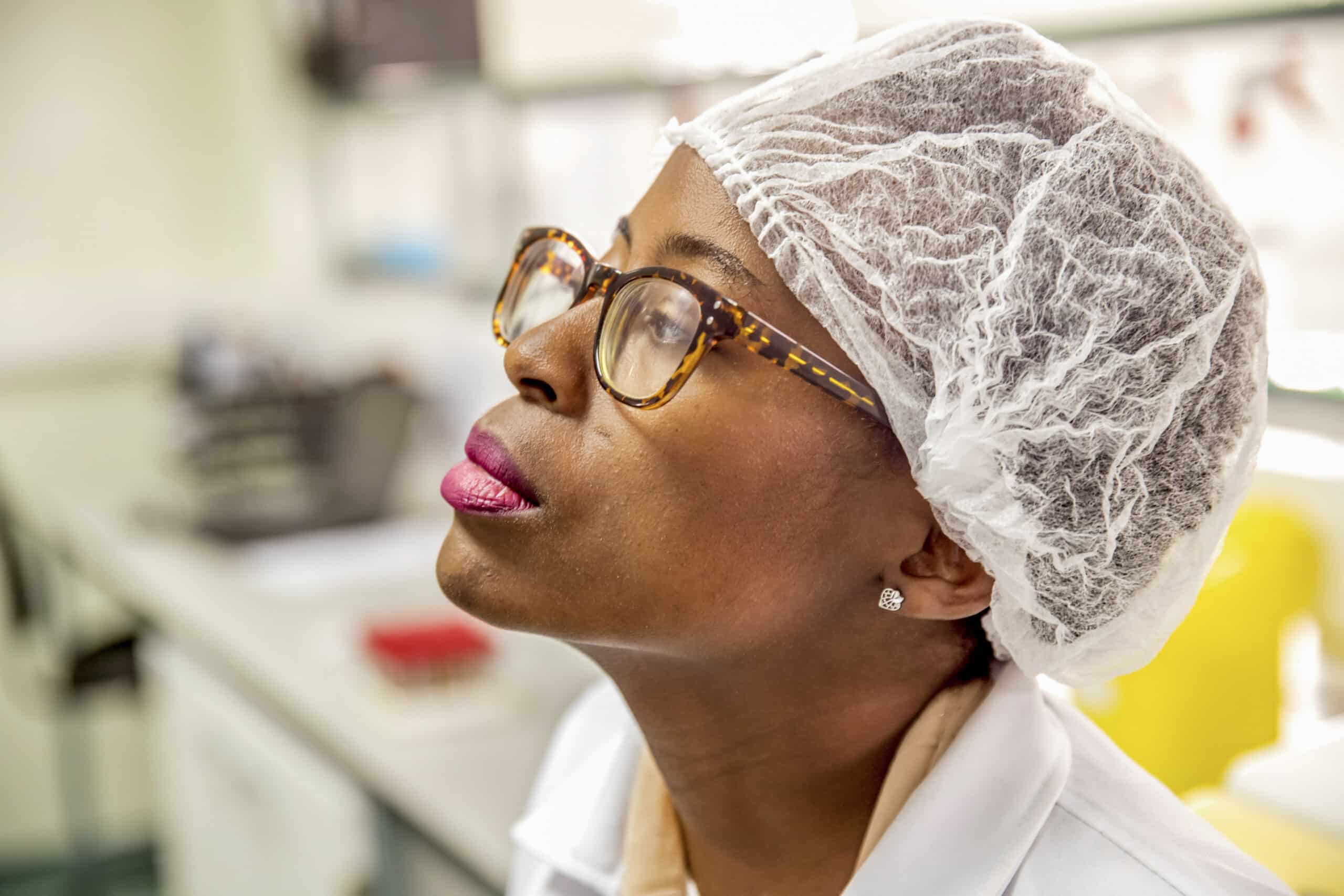 Afroamerikanische Wissenschaftlerin mit Brille und Haarnetz blickt in einer Laborumgebung nachdenklich nach oben. © Fotografie Tomas Rodriguez