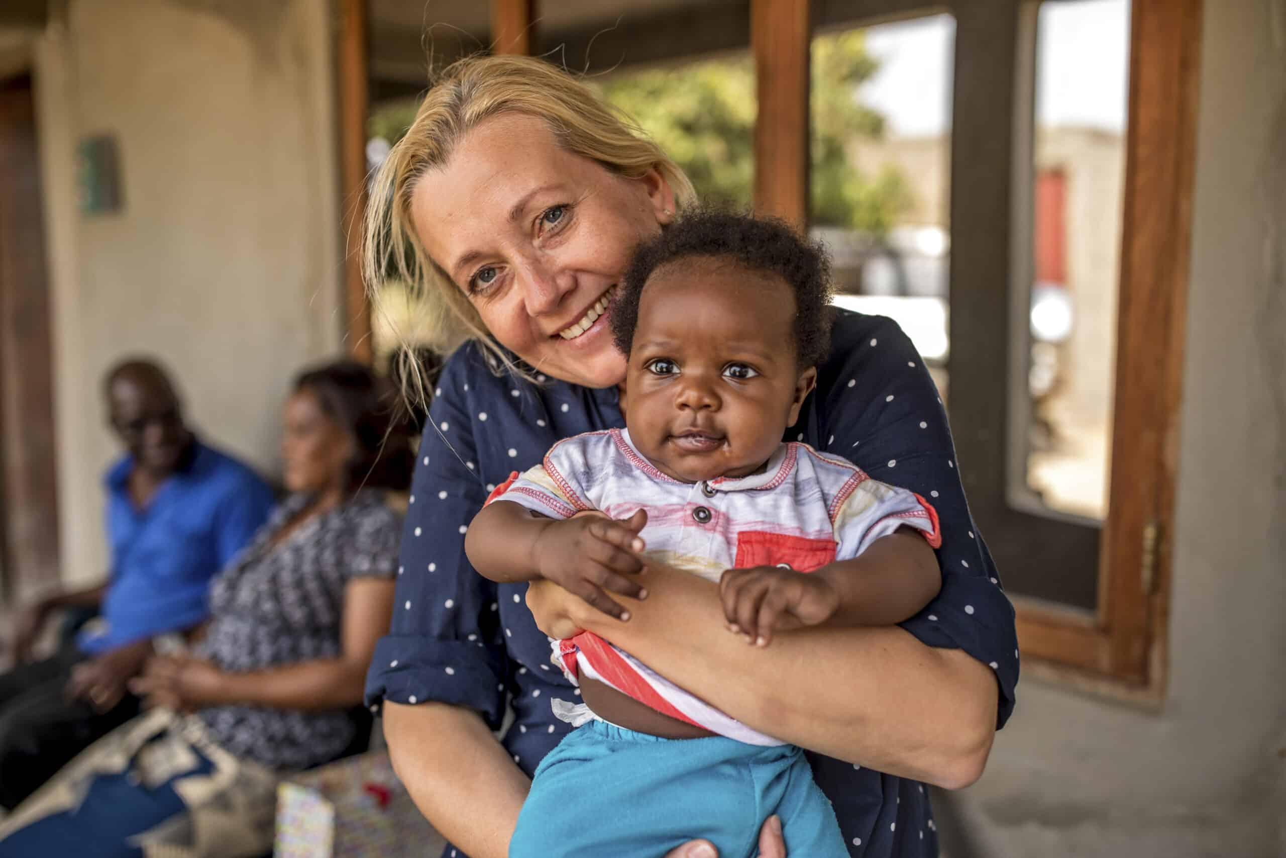 Eine Frau mit blondem Haar lächelt und hält ein Baby mit dunklem Haar im Arm. Beide sitzen auf einer Veranda, im Hintergrund sind Menschen zu sehen. © Fotografie Tomas Rodriguez