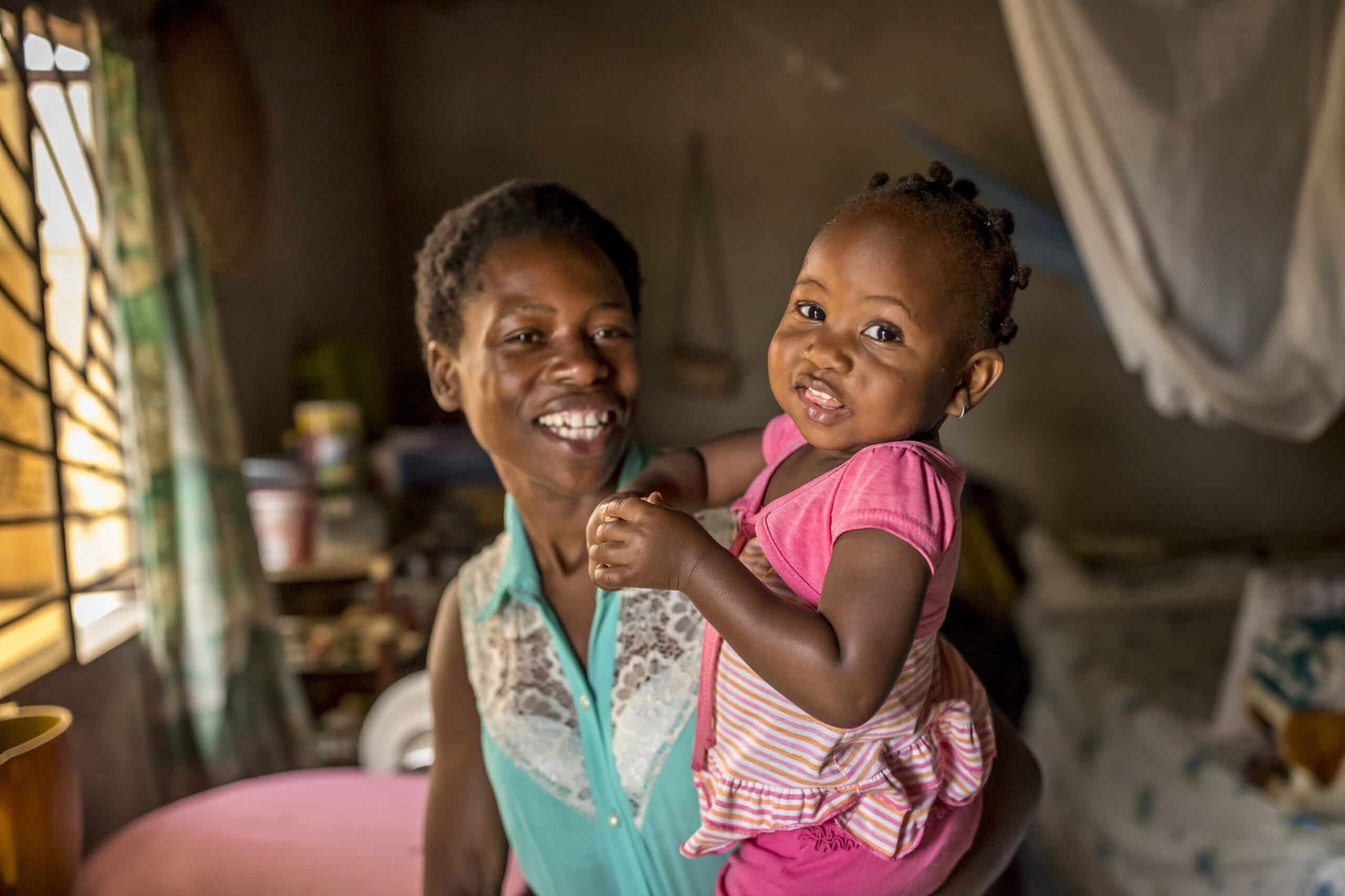 Eine fröhliche Mutter hält ihre kleine Tochter in einem sonnendurchfluteten Raum. Beide lächeln in die Kamera, während durch ein Fenster auf der linken Seite warmes Licht einfällt. © Fotografie Tomas Rodriguez