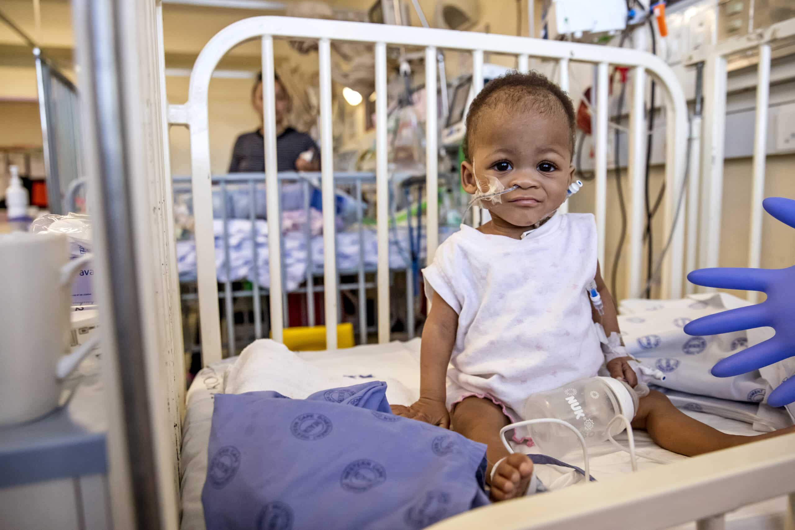 Ein Kleinkind mit einem kleinen Verband auf der Wange sitzt in einem Krankenhausbettchen und hält eine Tasse. Im Hintergrund ist ein weiteres Kind zu sehen. © Fotografie Tomas Rodriguez