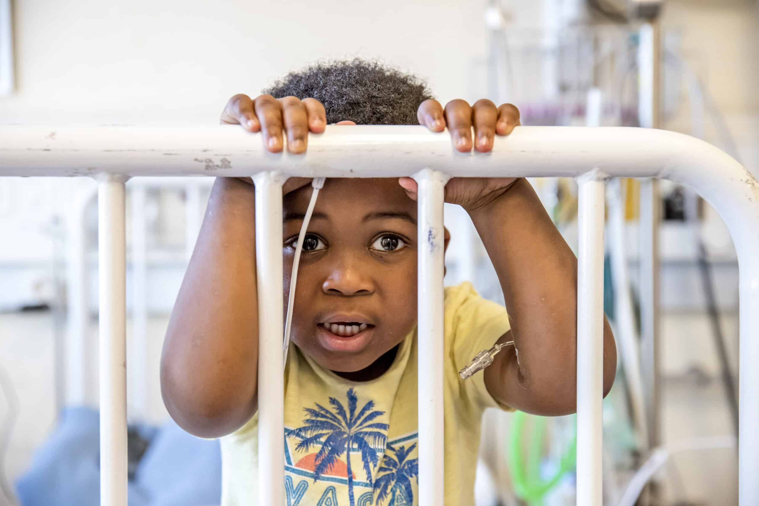Ein kleines Kind späht mit großen, neugierigen Augen und leicht geöffnetem Mund durch die Gitterstäbe eines weißen Kinderbetts in einem hell erleuchteten Raum und umklammert mit den Händen die Gitterstäbe. © Fotografie Tomas Rodriguez