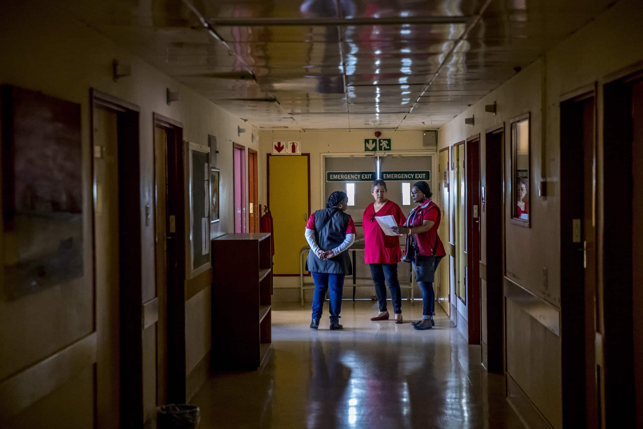 Drei medizinische Mitarbeiter besprechen Dokumente in einem Krankenhausflur bei schwacher Beleuchtung und sichtbaren Notausgangsschildern. © Fotografie Tomas Rodriguez