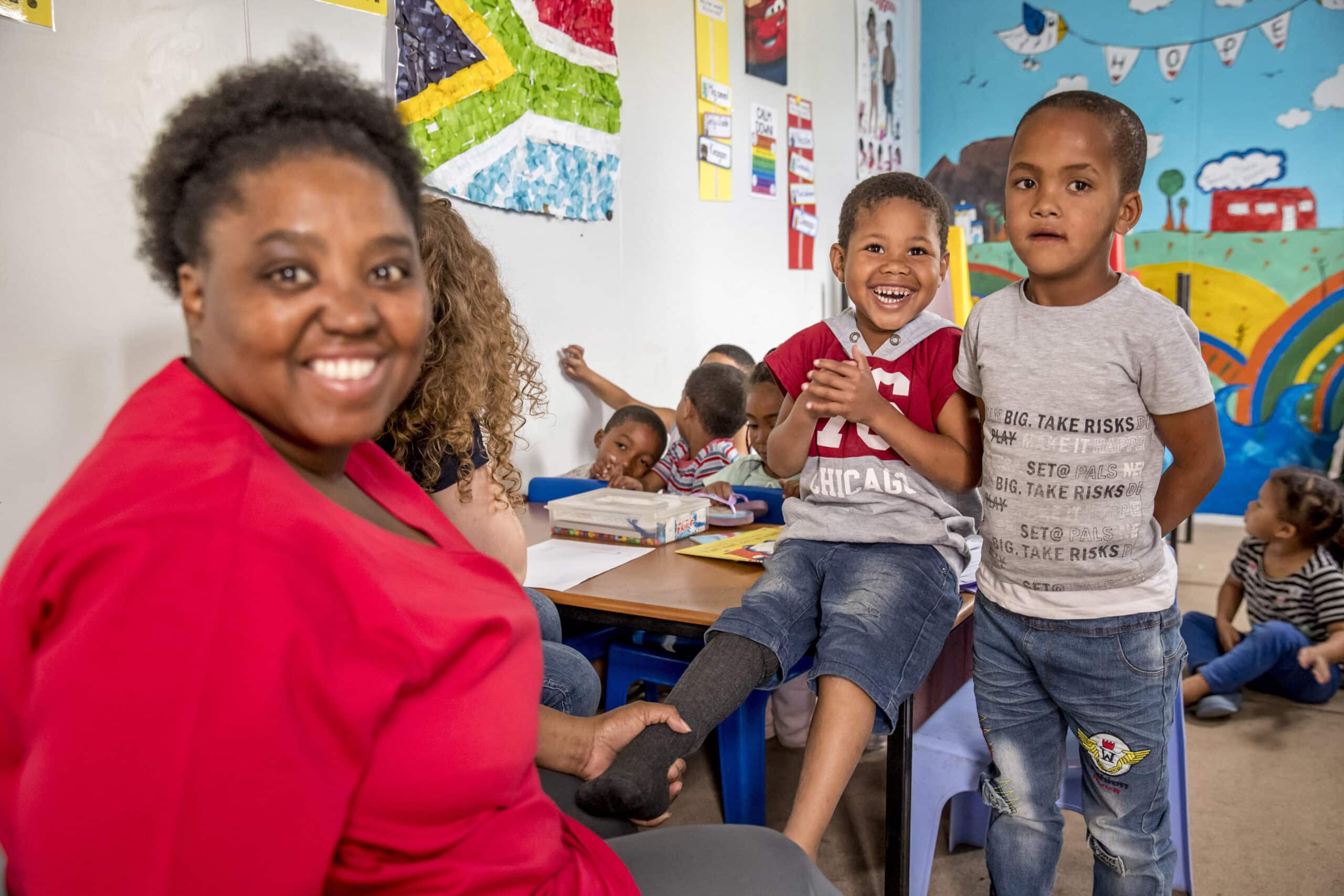 Eine fröhliche Klassenzimmerszene mit einer lächelnden Lehrerin und zwei glücklichen Jungen im Vordergrund, umgeben von anderen Kindern, die mit Aktivitäten beschäftigt sind, in einem farbenfrohen Raum, der mit Kinderkunstwerken geschmückt ist. © Fotografie Tomas Rodriguez
