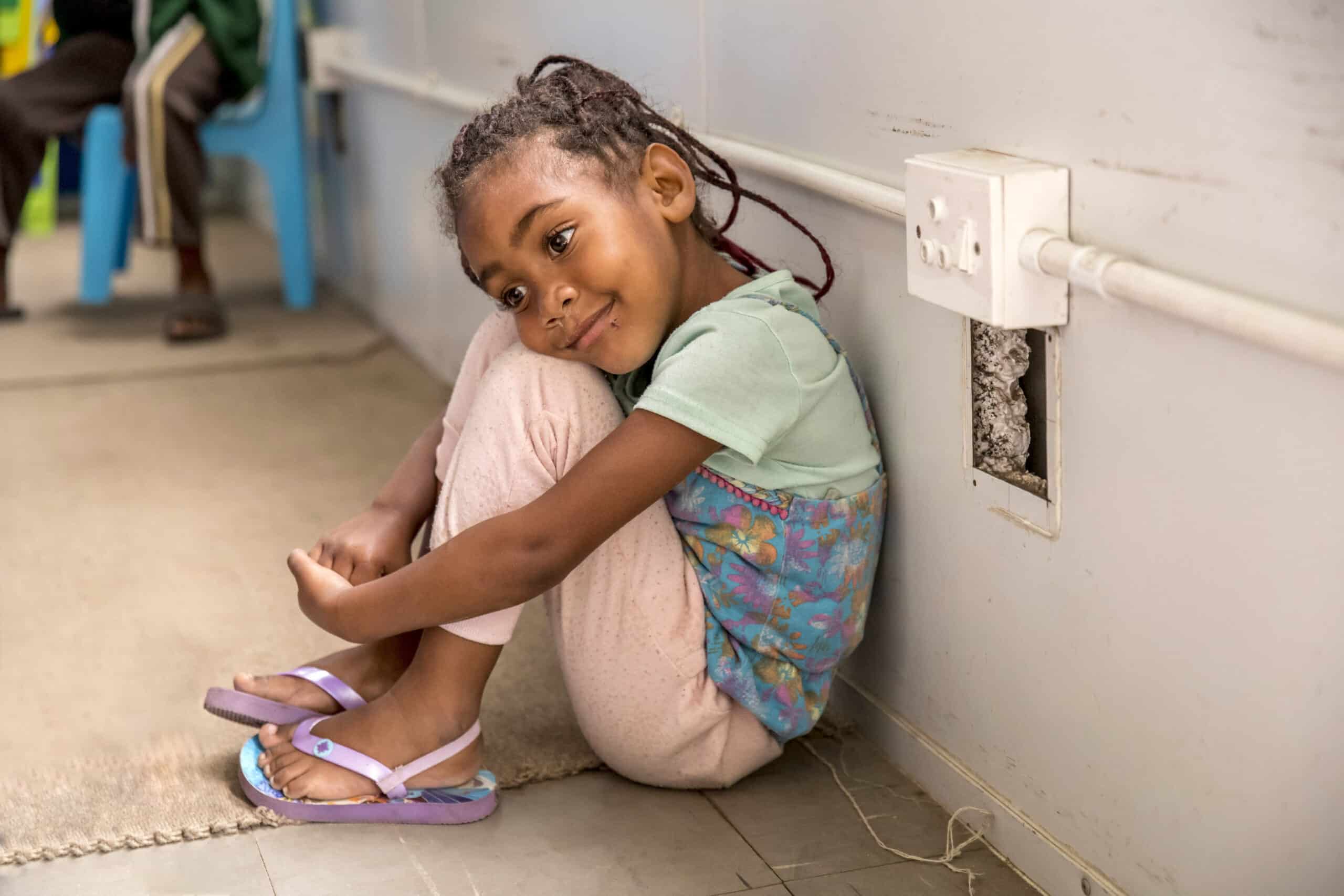 Ein junges Mädchen sitzt auf dem Boden, lehnt an einer weißen Wand und lächelt seitlich in die Kamera. Es trägt Flip-Flops und ein buntes Oberteil. © Fotografie Tomas Rodriguez