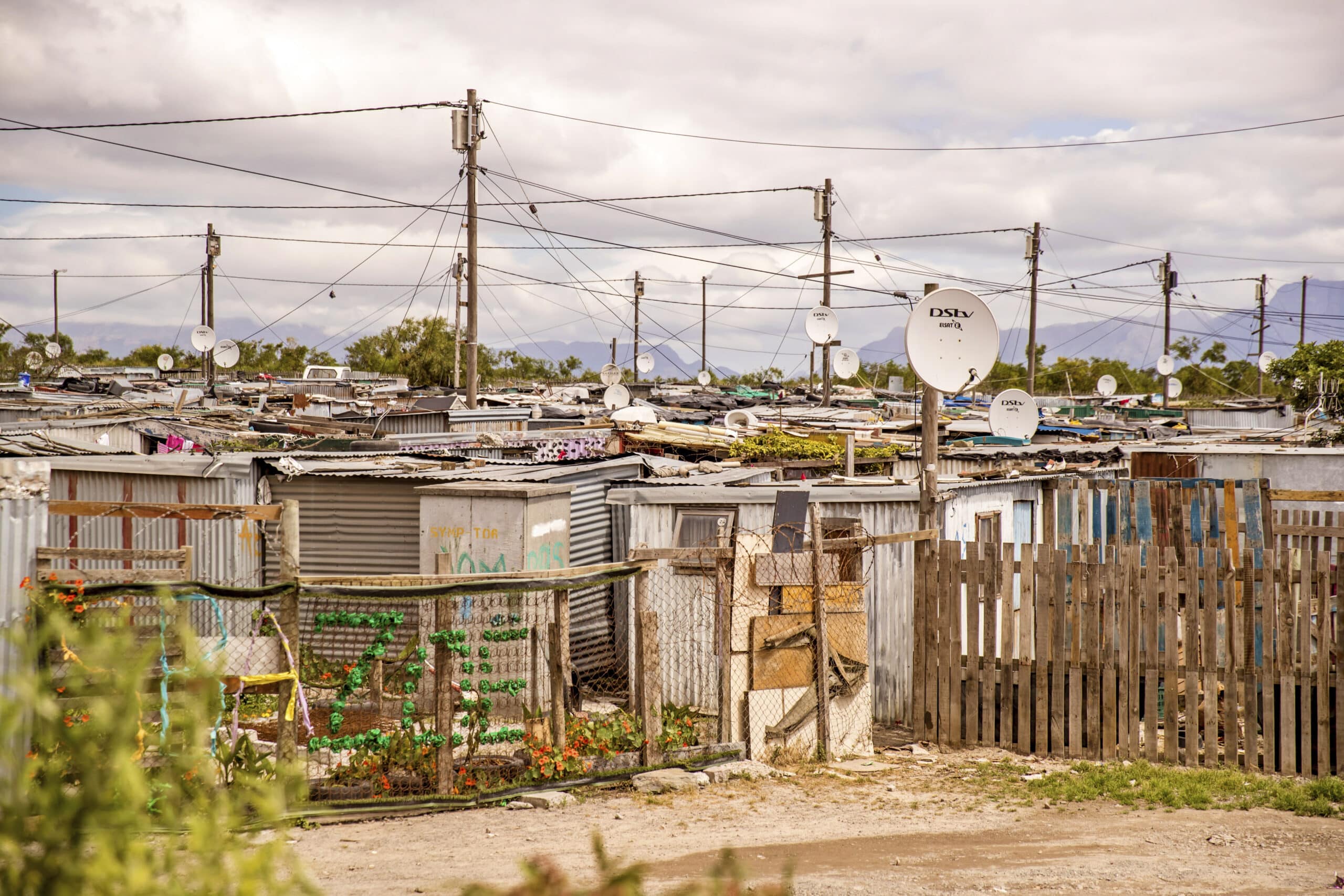 Eine provisorische Siedlung mit dicht gedrängten Hütten mit Wellblechdächern, umgeben von Zäunen; viele Satellitenschüsseln sind vor einem bewölkten Himmel sichtbar. © Fotografie Tomas Rodriguez