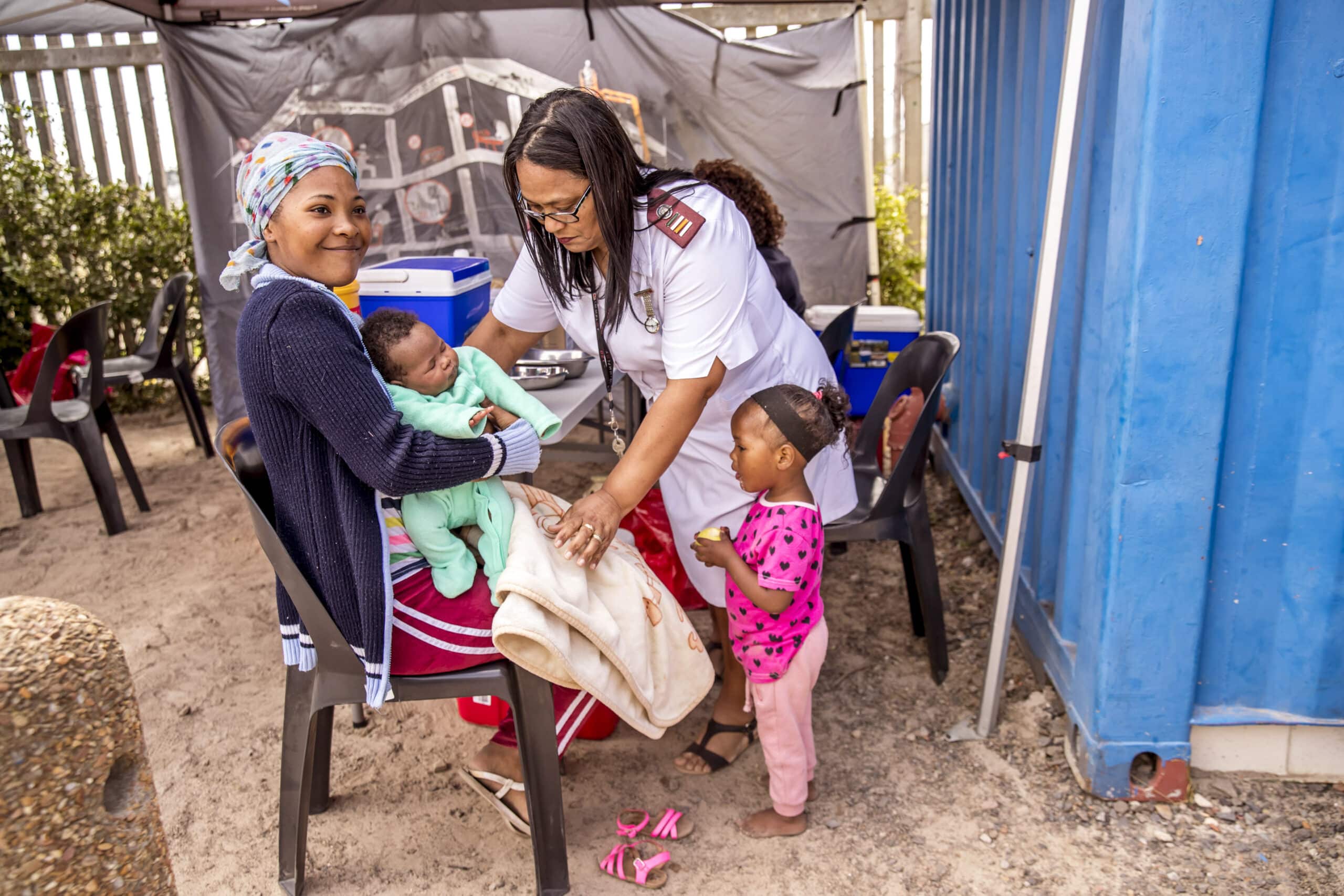 Eine Mitarbeiterin des Gesundheitswesens interagiert mit einer Mutter, die ein Baby und ein kleines Mädchen hält, in einer provisorischen Klinik im Freien, umgeben von medizinischem Material und einem blauen Zelt. © Fotografie Tomas Rodriguez