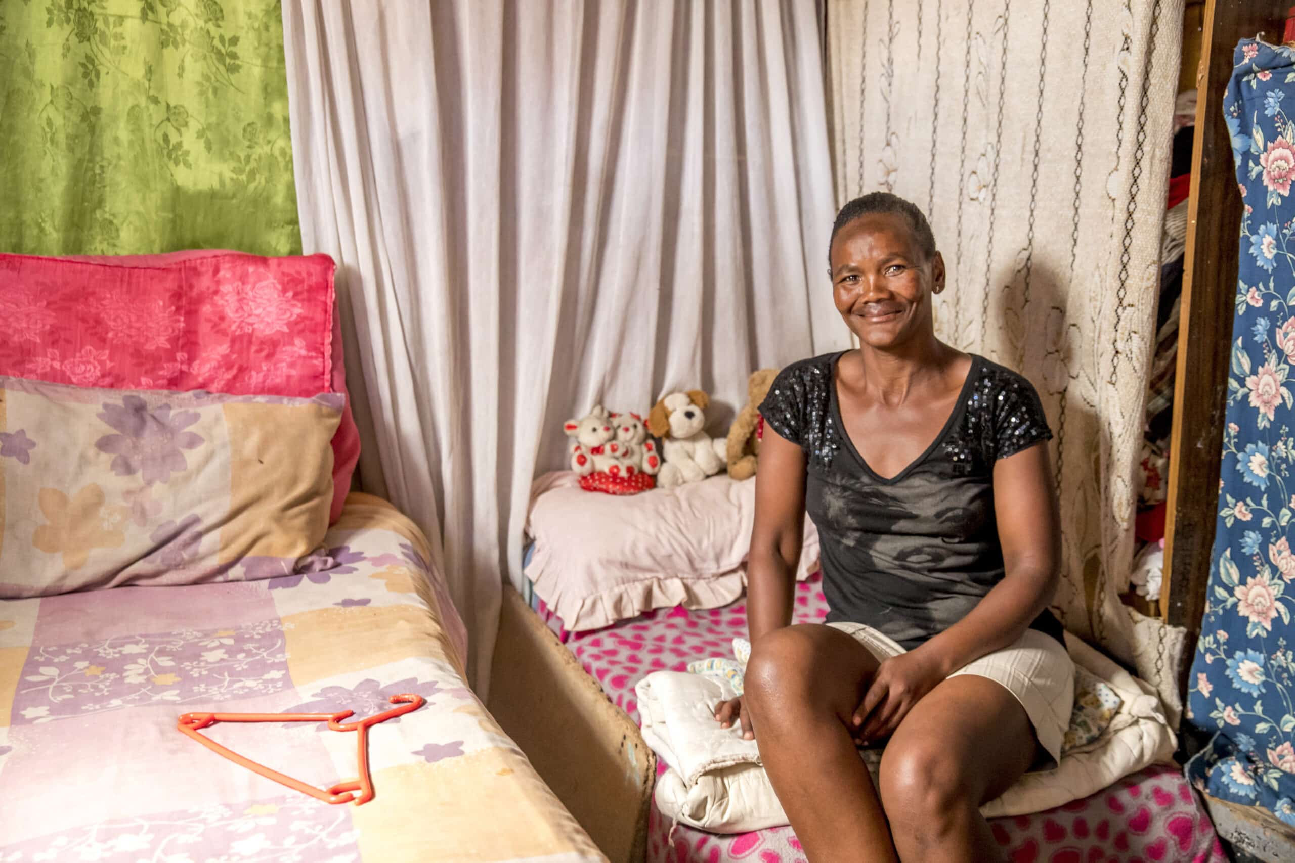 Eine Frau sitzt auf einem Bett in einem einfachen Zimmer mit bunten Vorhängen und Bettwäsche und lächelt in die Kamera. Ein kleiner Tisch und Teddybären sind ebenfalls zu sehen. © Fotografie Tomas Rodriguez