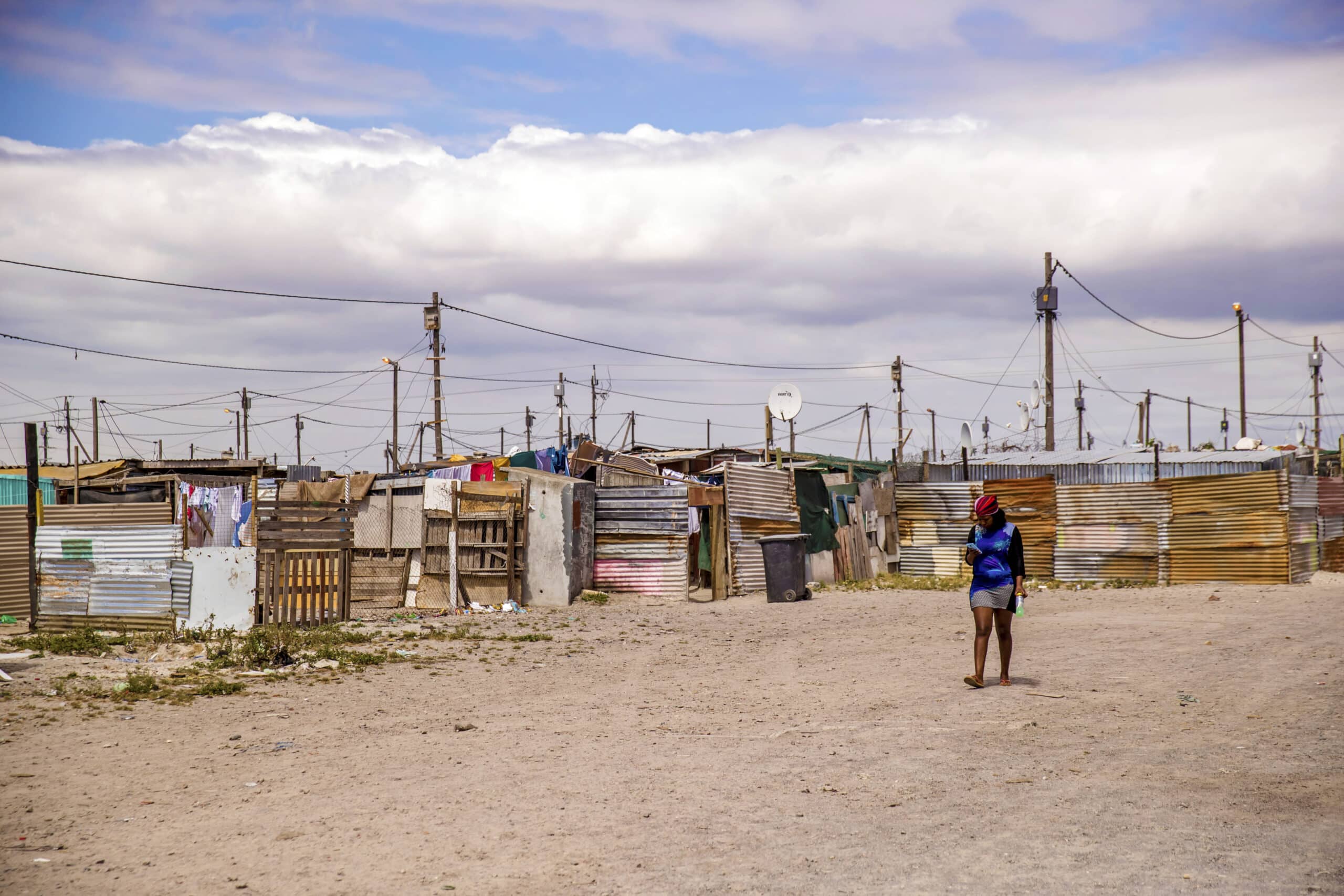 Eine Frau geht unter einem bewölkten Himmel durch eine weitläufige informelle Siedlung mit provisorischen Behausungen aus Wellblech und anderen Materialien. Auf einigen Hütten sind Satellitenschüsseln zu sehen. © Fotografie Tomas Rodriguez