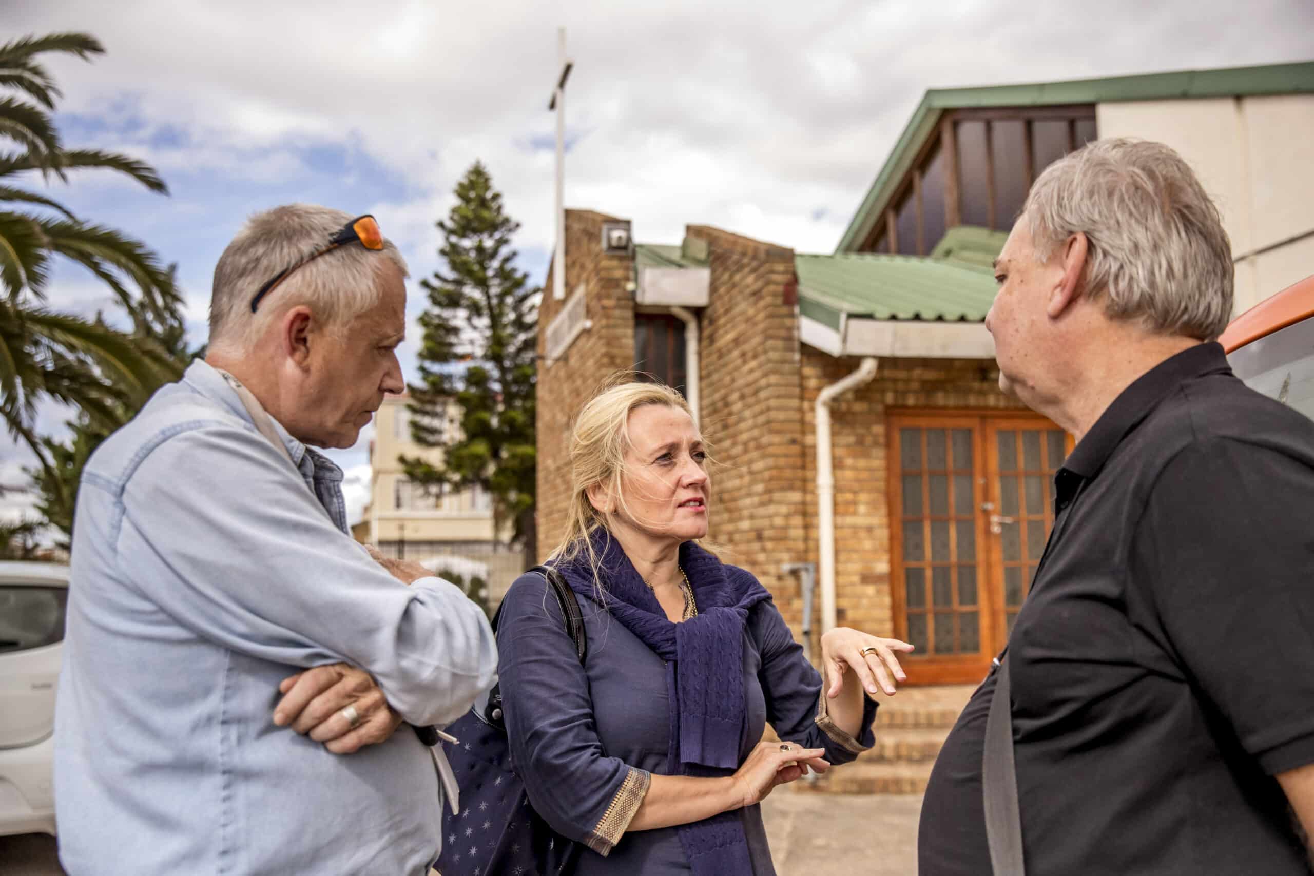 Zwei ältere Männer und eine Frau unterhalten sich im Freien in der Nähe eines Gebäudes unter einem bewölkten Himmel. Die Frau, die in der Mitte steht, gestikuliert, während sie mit einem der Männer spricht. © Fotografie Tomas Rodriguez