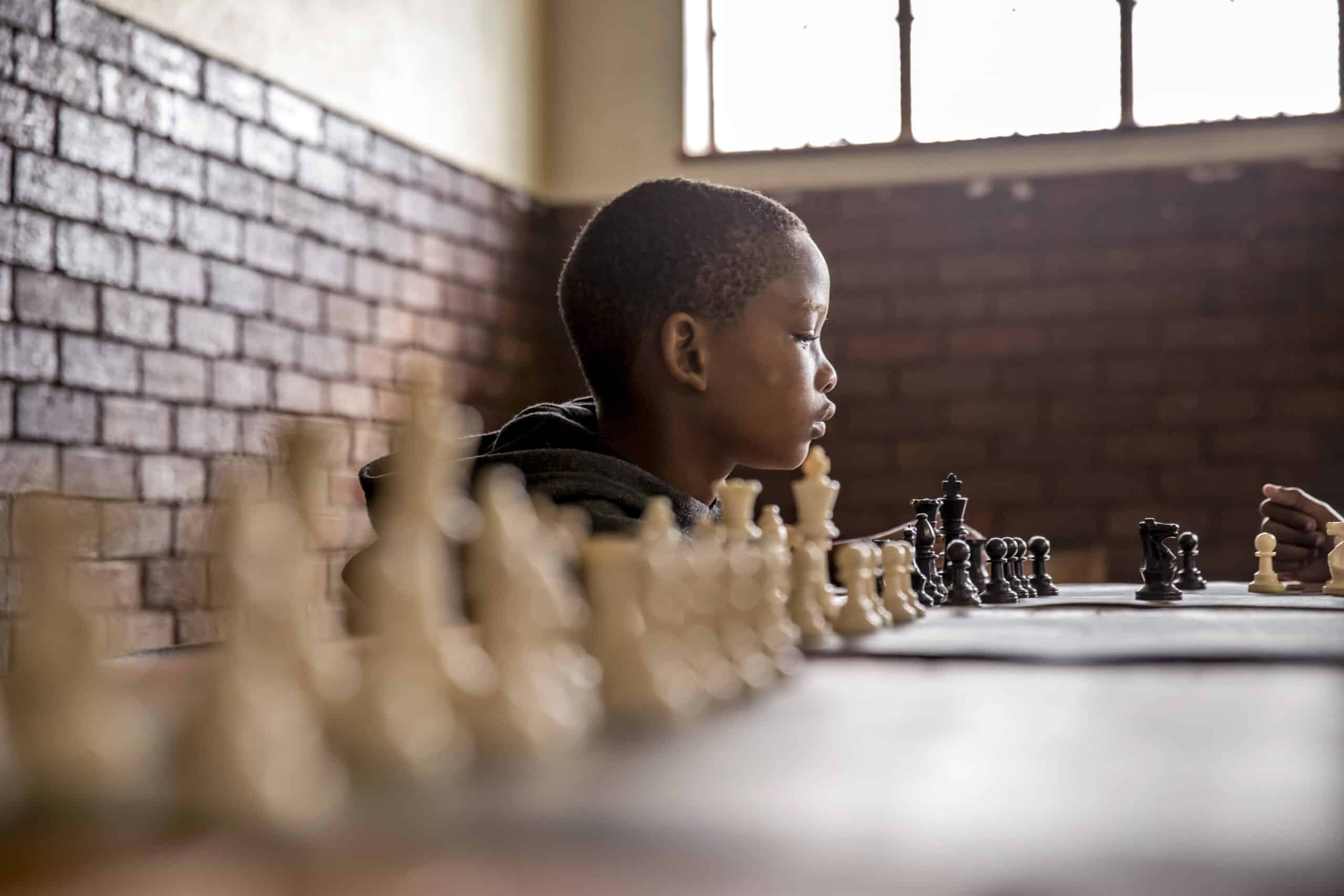 Ein junger Schwarzer konzentriert sich tief auf ein Schachspiel und betrachtet das Schachbrett aus nächster Nähe in einem Raum mit Backsteinwänden und großen Fenstern. © Fotografie Tomas Rodriguez