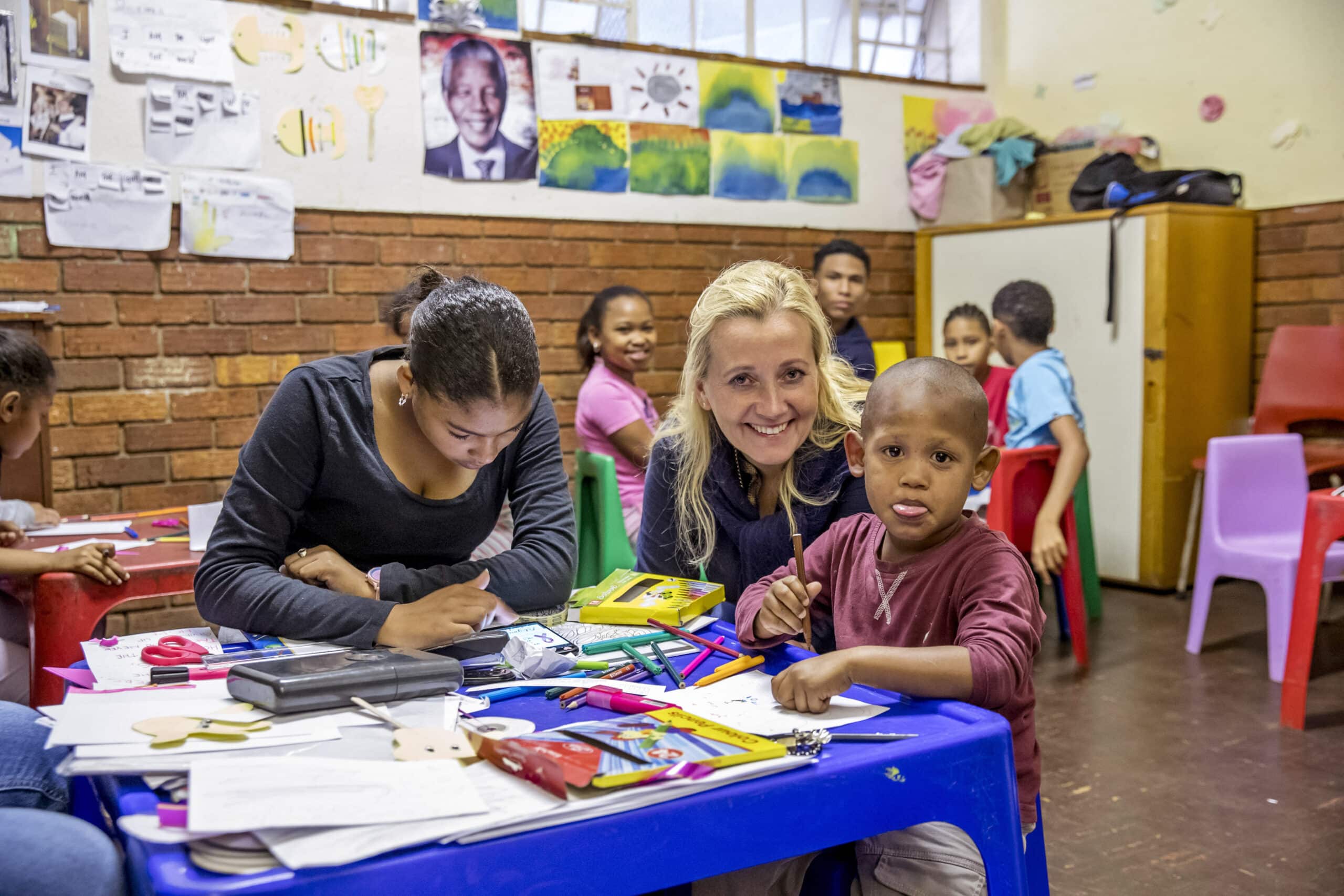 Eine Lehrerin lächelt in einem Klassenzimmer mit unterschiedlichen Schülern in die Kamera, von denen einige an Tischen voller bunter Schulsachen malen. © Fotografie Tomas Rodriguez