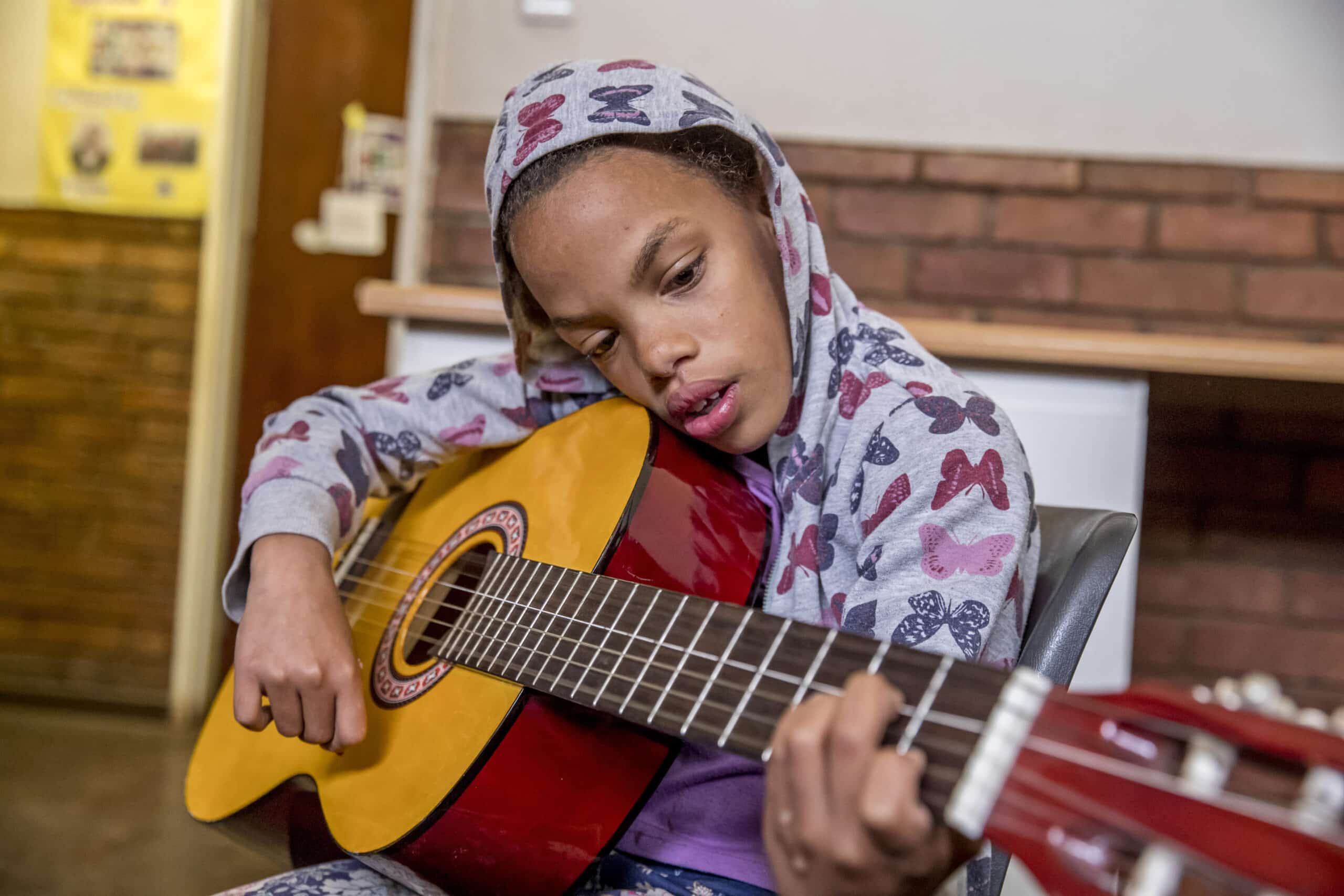 Ein junges Mädchen mit Kapuzenpullover und gemusterten Leggings sitzt im Innenbereich und spielt konzentriert Akustikgitarre. An der Wand hinter ihr hängen Informationsplakate. © Fotografie Tomas Rodriguez