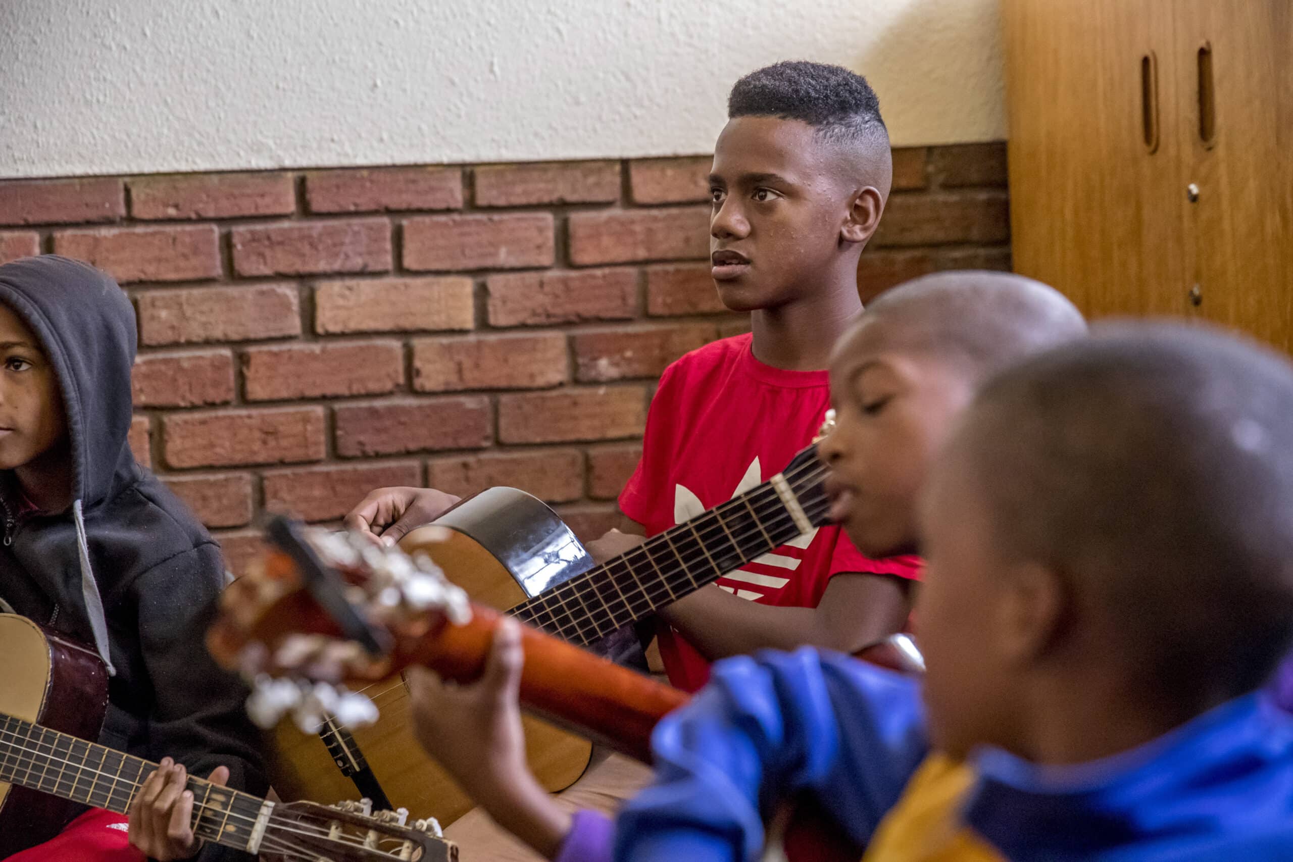Ein Teenager im roten Hemd blickt nachdenklich zur Seite und hält eine Gitarre in der Hand. In einem Raum mit Backsteinwänden sind andere kleine Kinder um ihn herum, die ebenfalls Gitarre spielen. © Fotografie Tomas Rodriguez