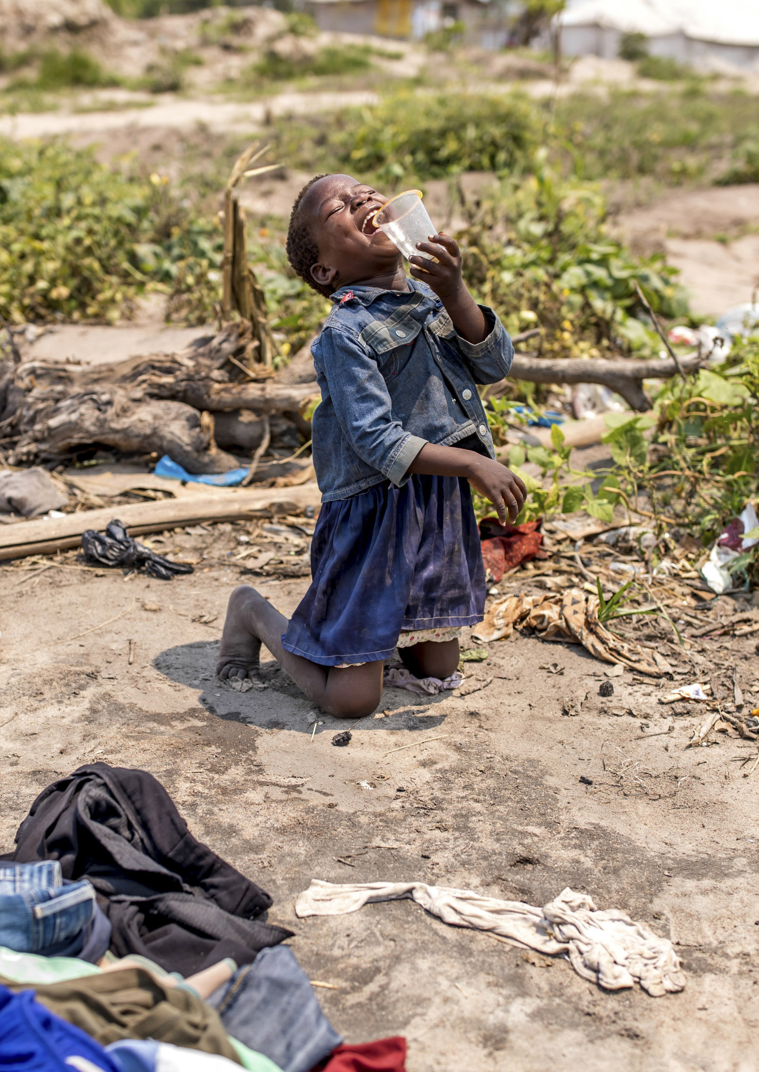 Ein kleiner Junge kniet auf dem Boden und trinkt aus einer Tasse, umgeben von verstreuter Kleidung und Abfall. Er bringt damit seine starken Emotionen in einer vernachlässigten Umgebung zum Ausdruck. © Fotografie Tomas Rodriguez