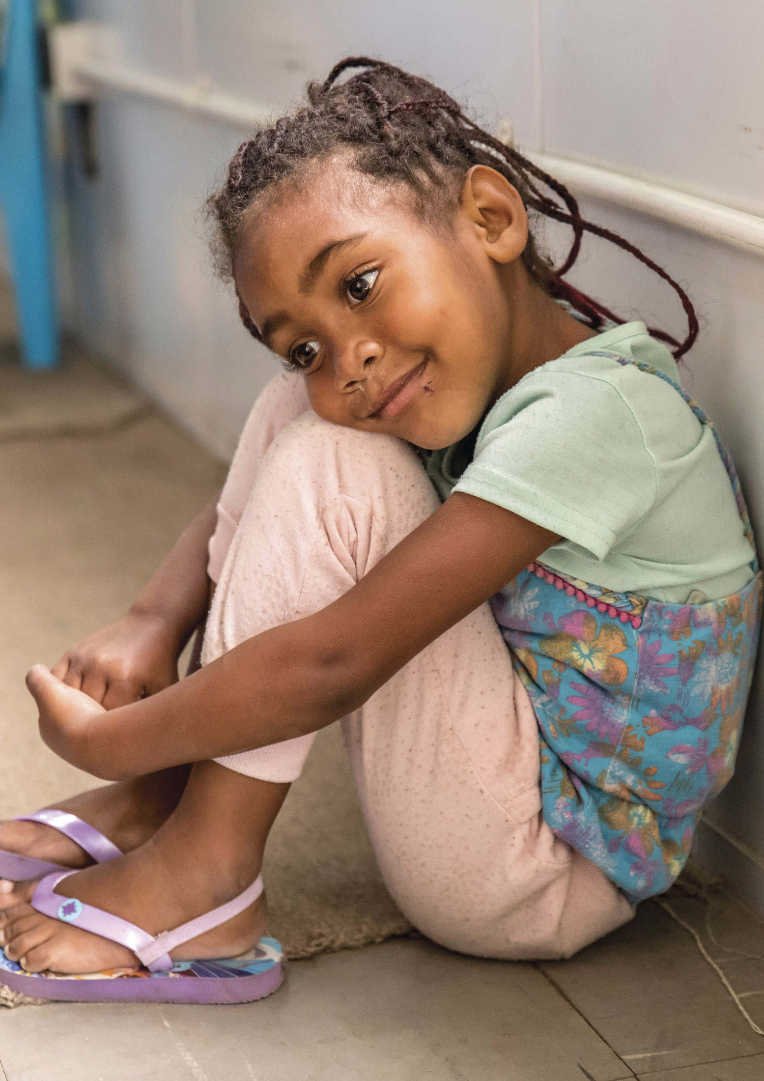 Ein junges Mädchen mit geflochtenen Haaren sitzt mit angezogenen Beinen auf dem Boden, lehnt an einer Wand, trägt ein pastellfarbenes T-Shirt und einen gemusterten Rucksack und blickt nachdenklich weg. © Fotografie Tomas Rodriguez