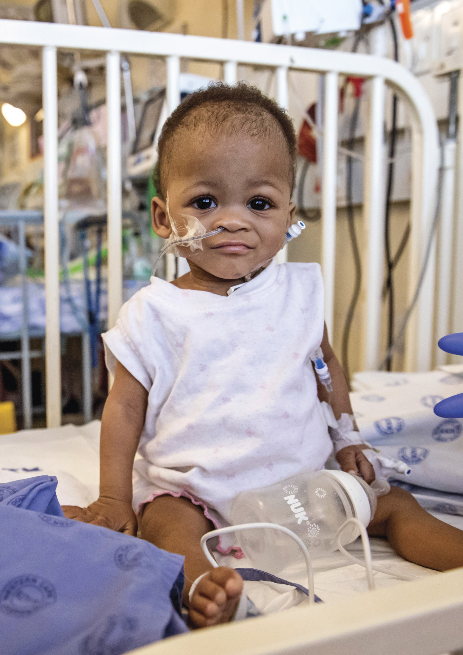 Ein Kleinkind mit Nasensonden sitzt auf einem Krankenhausbett, trägt ein weißes Hemd und blickt mit einem leichten Lächeln in die Kamera. In der Umgebung sind medizinische Geräte und Bettzeug zu sehen. © Fotografie Tomas Rodriguez