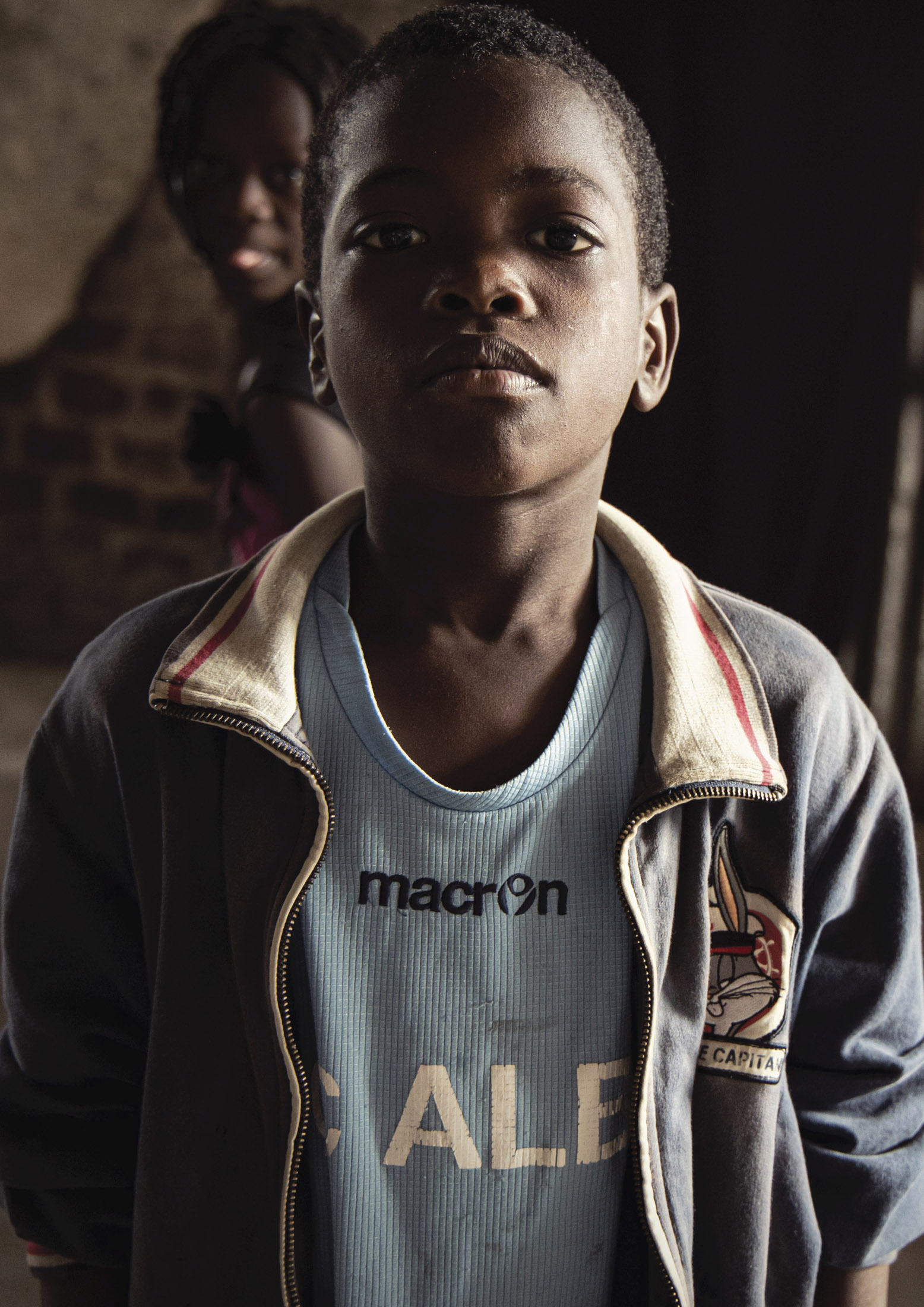 Ein Porträt eines kleinen Jungen mit ernstem Gesichtsausdruck, der ein Sporttrikot und eine Jacke trägt. Er steht in einem Innenbereich mit sanfter Beleuchtung, im Hintergrund ist ein lächelndes Mädchen zu sehen. © Fotografie Tomas Rodriguez