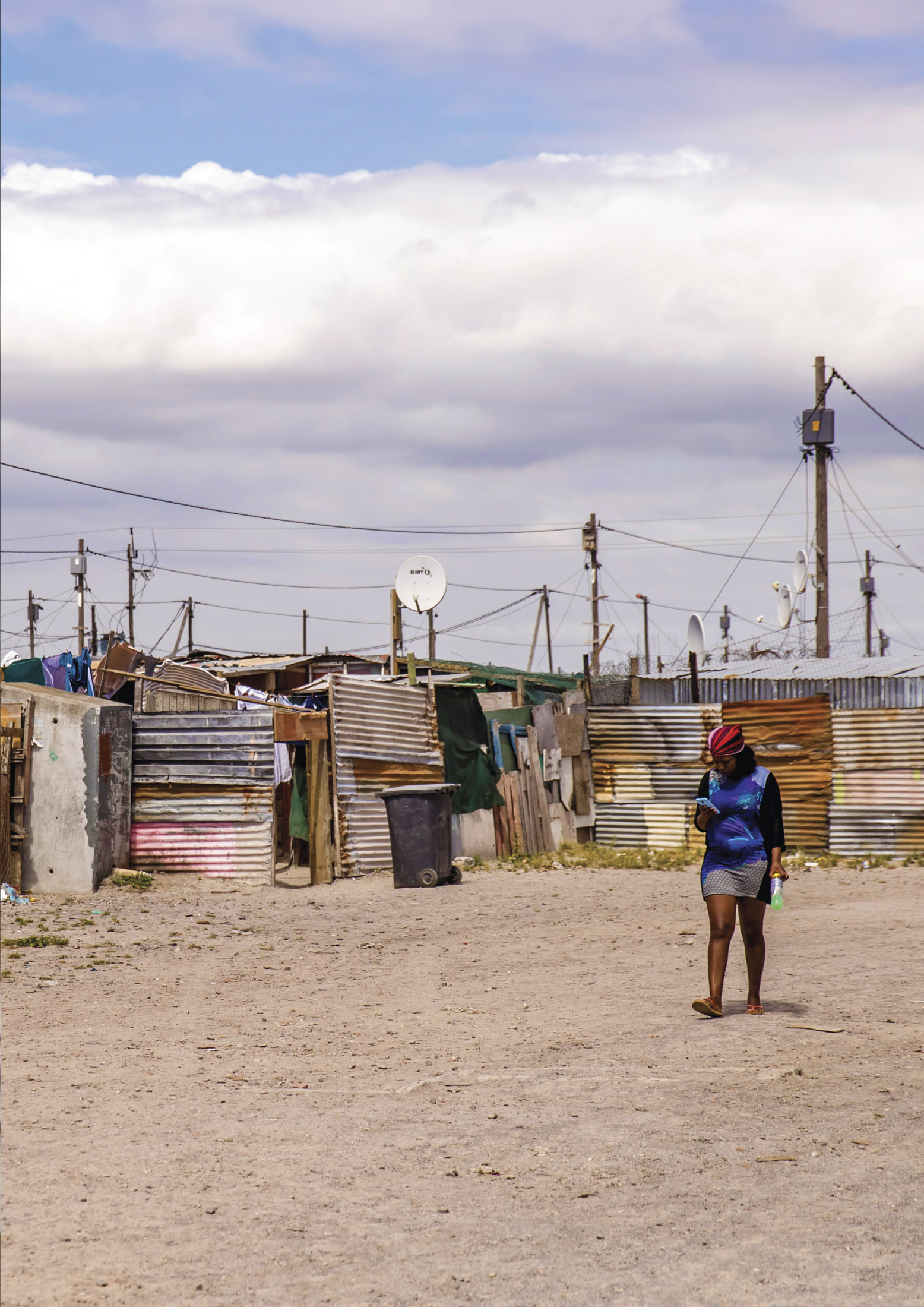 Eine Frau geht von der Kamera weg eine staubige Straße entlang, die in einer dünn besiedelten Gegend unter einem bewölkten Himmel von provisorischen Behausungen aus Wellblech gesäumt ist. Satellitenschüsseln sind zu sehen. © Fotografie Tomas Rodriguez