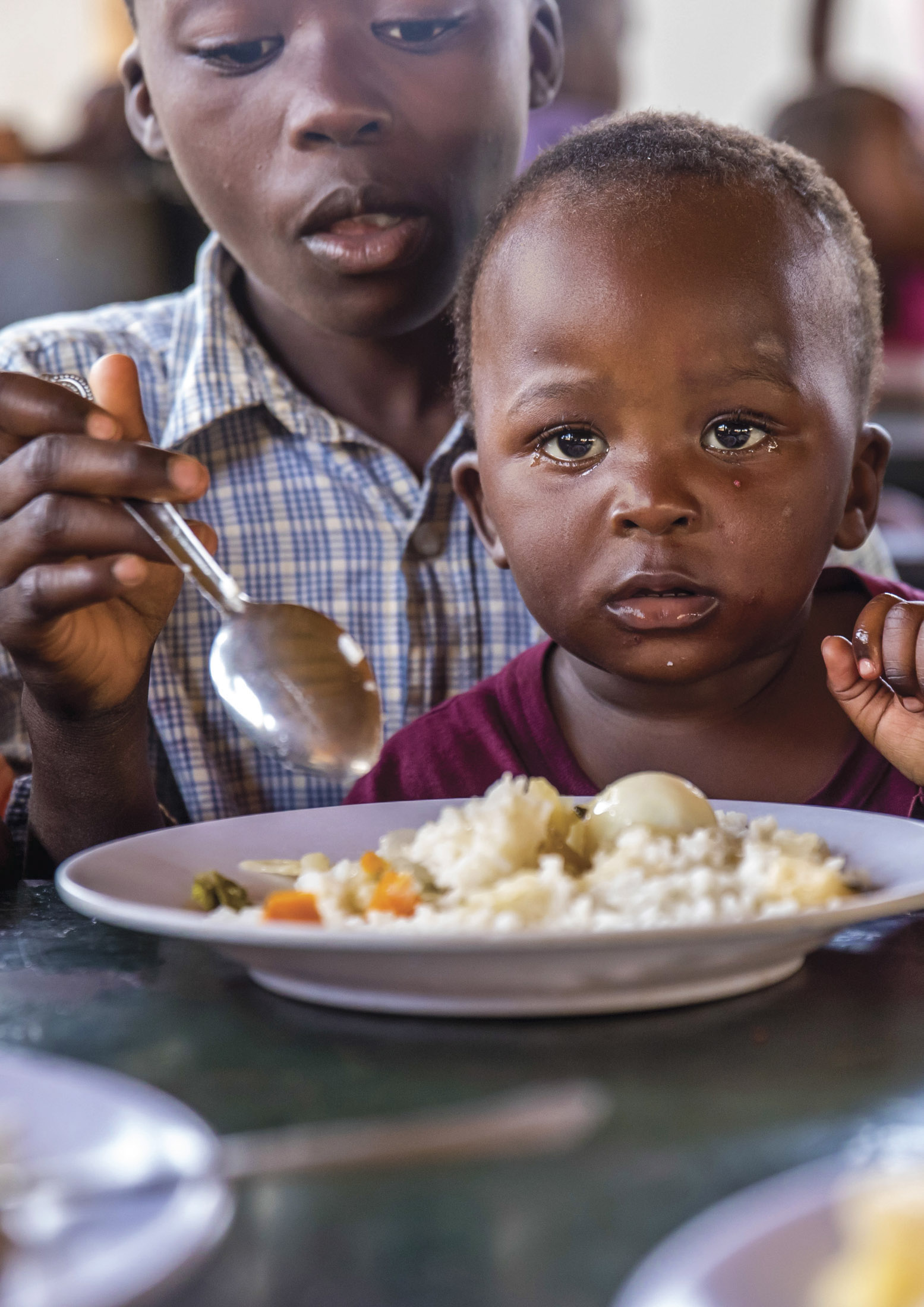 Ein kleines Kind mit ernster Miene sitzt an einem Tisch mit einem Teller Reis und Gemüse. Ein älteres Kind daneben hält einen Löffel und ist bereit zu essen. © Fotografie Tomas Rodriguez