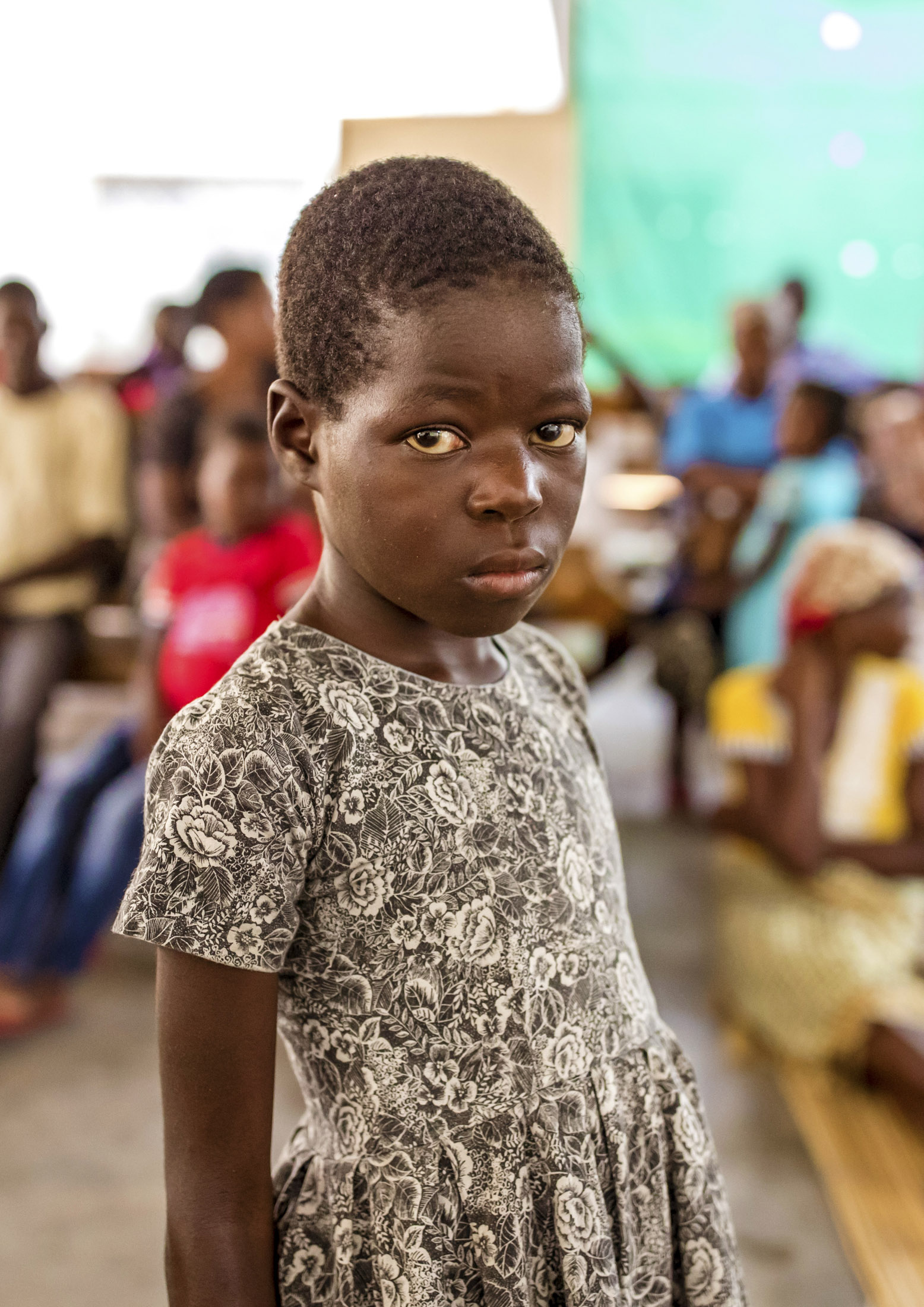 Ein junges Mädchen mit ernster Miene und einem geblümten Kleid steht in einem überfüllten Raum, möglicherweise bei einer Gemeinschaftsveranstaltung. © Fotografie Tomas Rodriguez