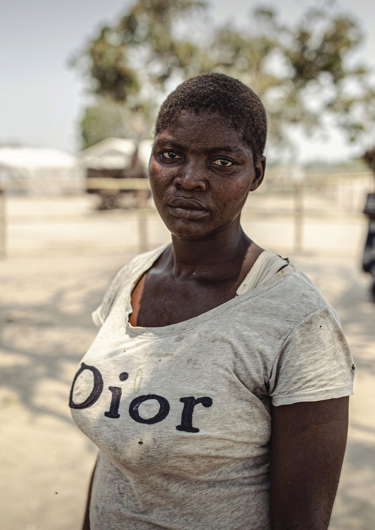 Ein Porträt einer jungen Frau mit ernster Miene, die im Freien steht. Sie trägt ein T-Shirt mit dem Aufdruck „Dior“ vor einem unscharfen Hintergrund mit Bäumen und Gebäuden. © Fotografie Tomas Rodriguez